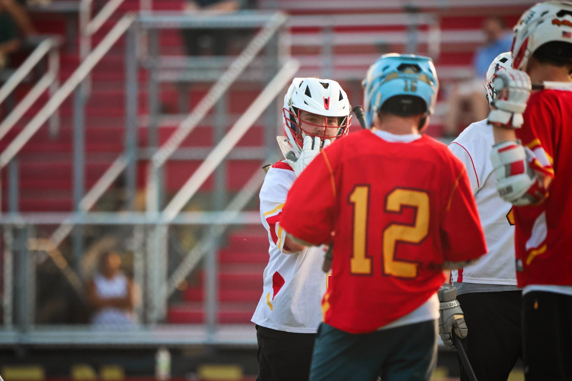 Lacrosse players in red and white uniforms stand on the field near bleachers, seen from behind and side view.