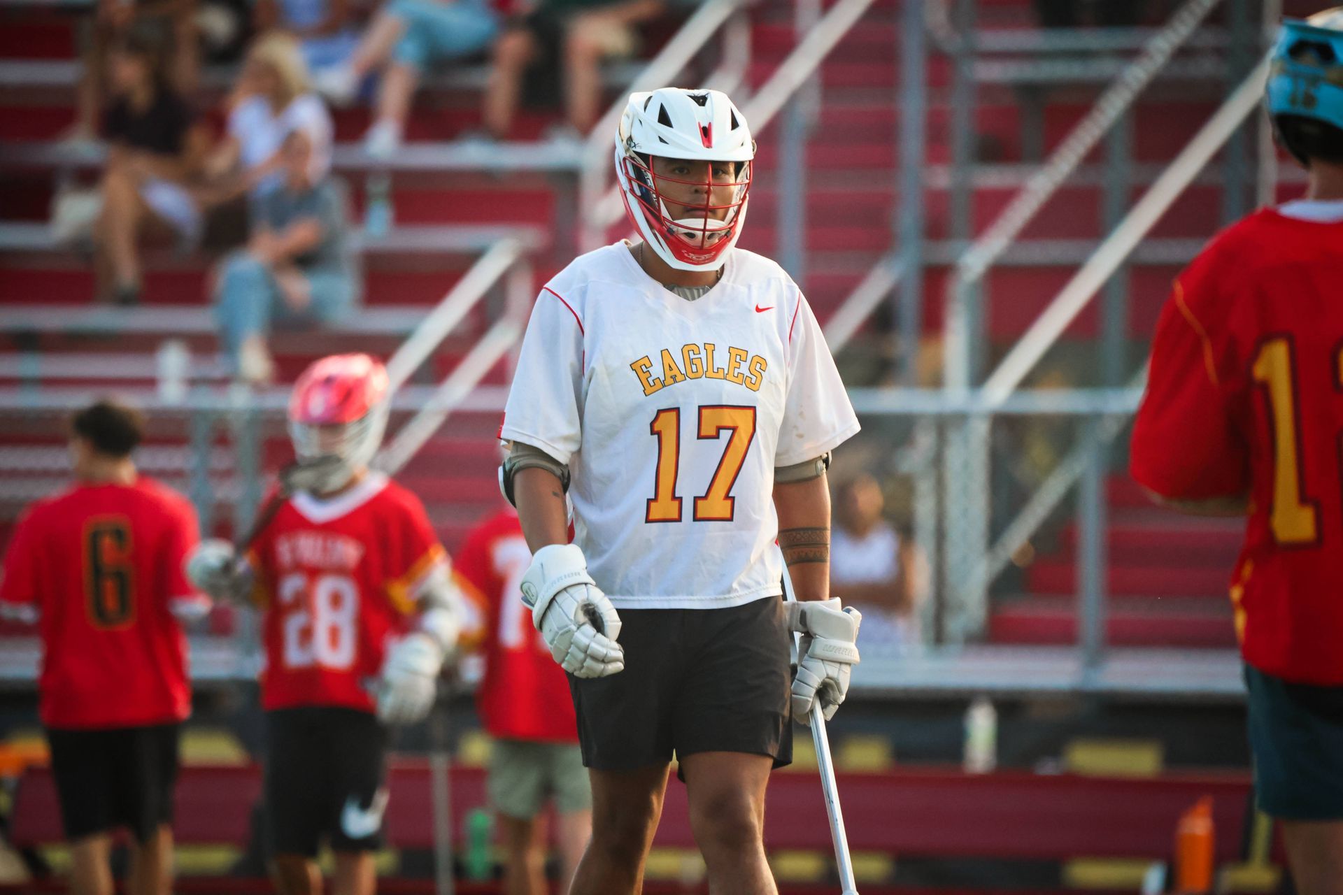 Lacrosse player in white jersey number 17 stands on the field with stick, teammates blurred behind him.