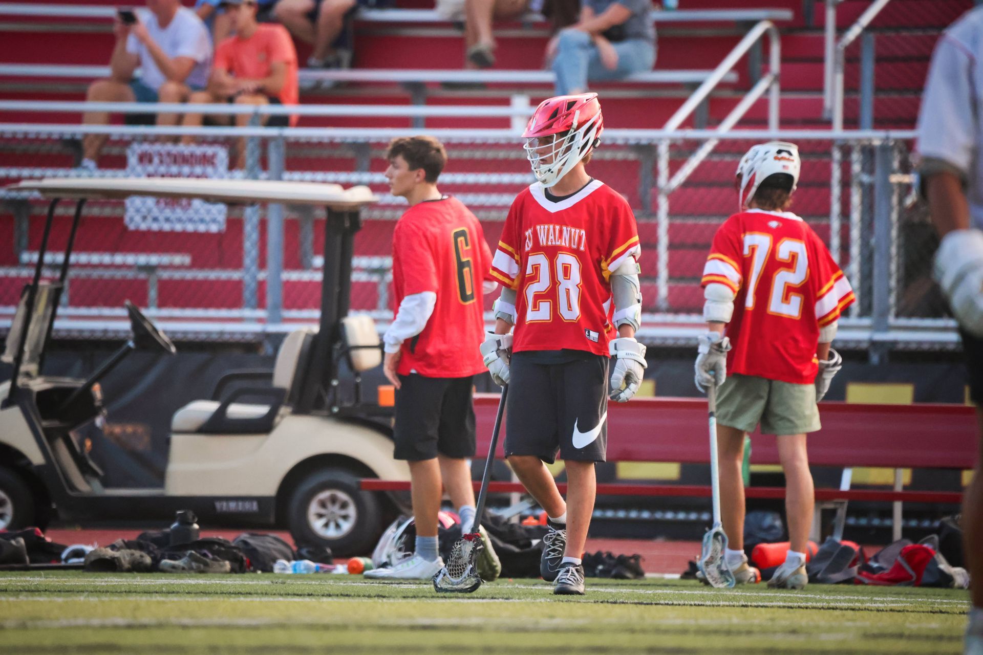 Three lacrosse players in red jerseys stand on the sideline near a golf cart at a field