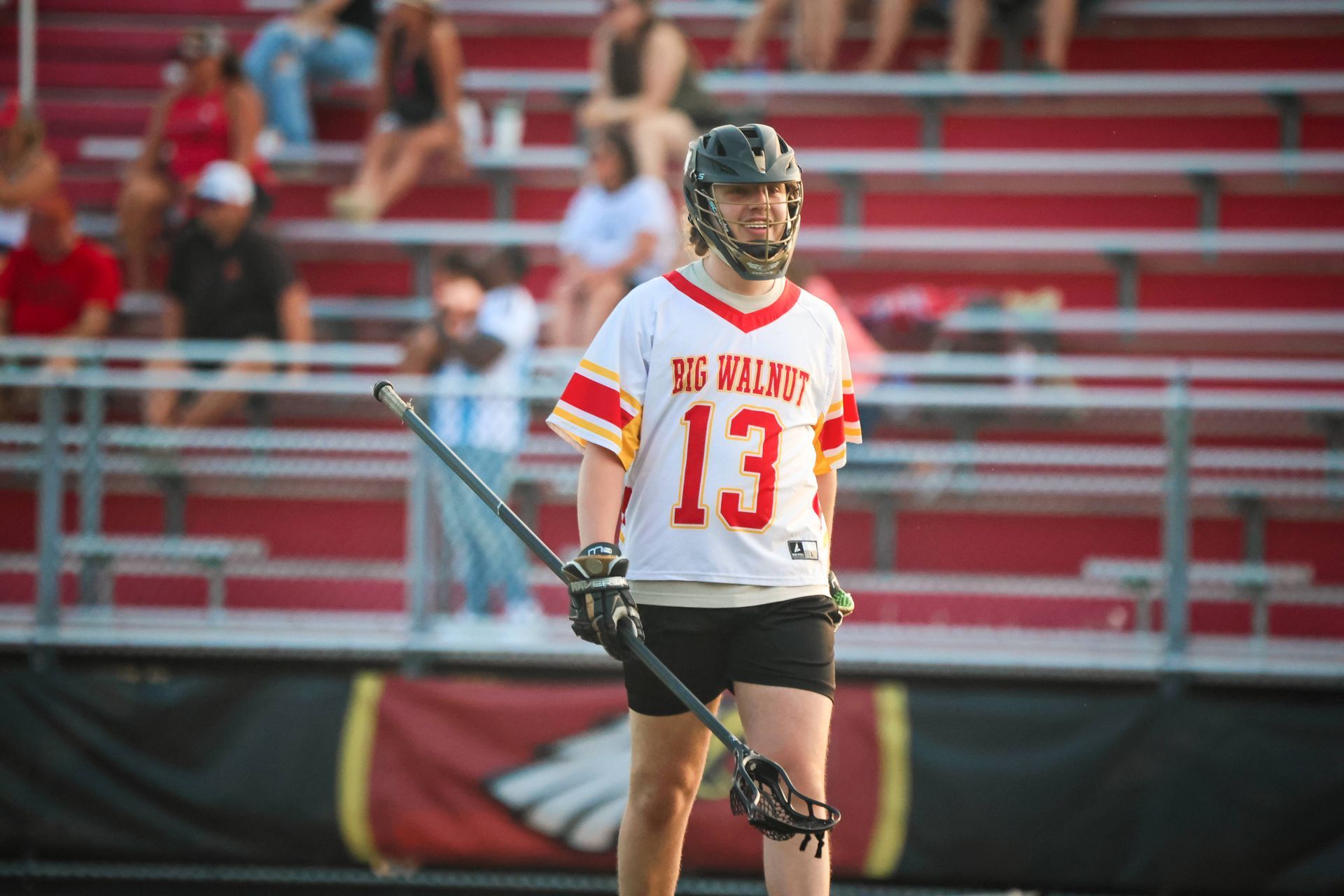 Lacrosse player in white and red jersey holding a stick on a field with stadium seating behind him