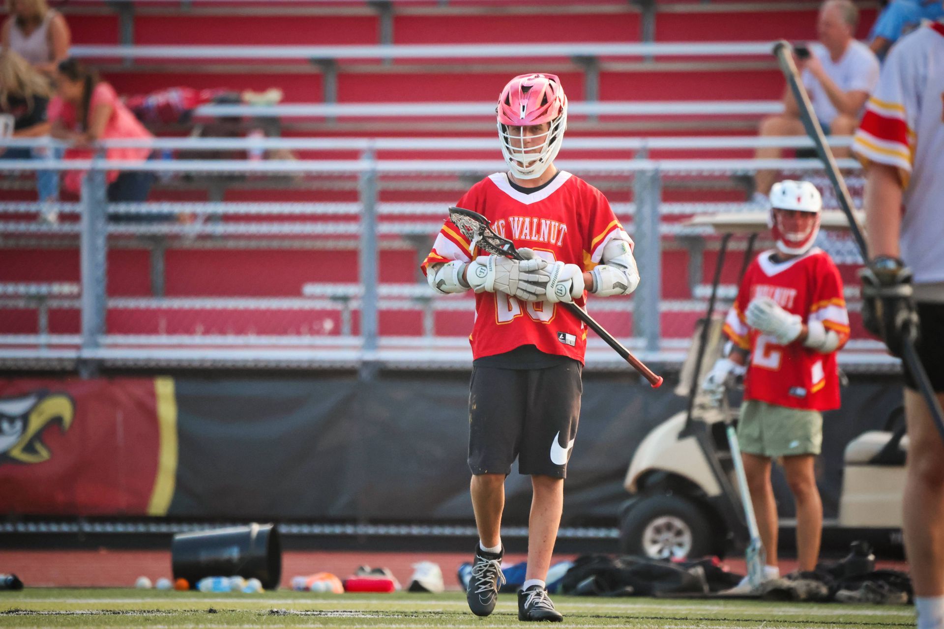 Lacrosse player in red jersey holding a stick on the field with bleachers in the background