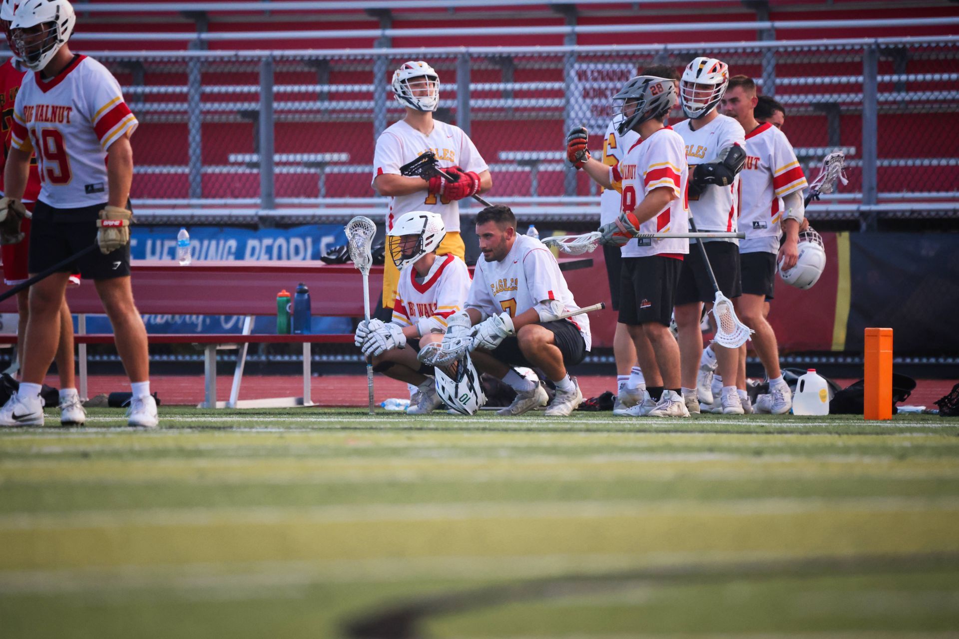 Lacrosse players gathered on the sideline during a game, with one kneeling and coaches nearby.