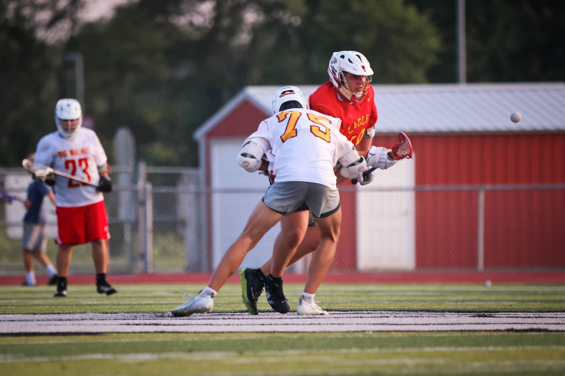 Two lacrosse players in red and white uniforms jostle for position on a field near a red building.