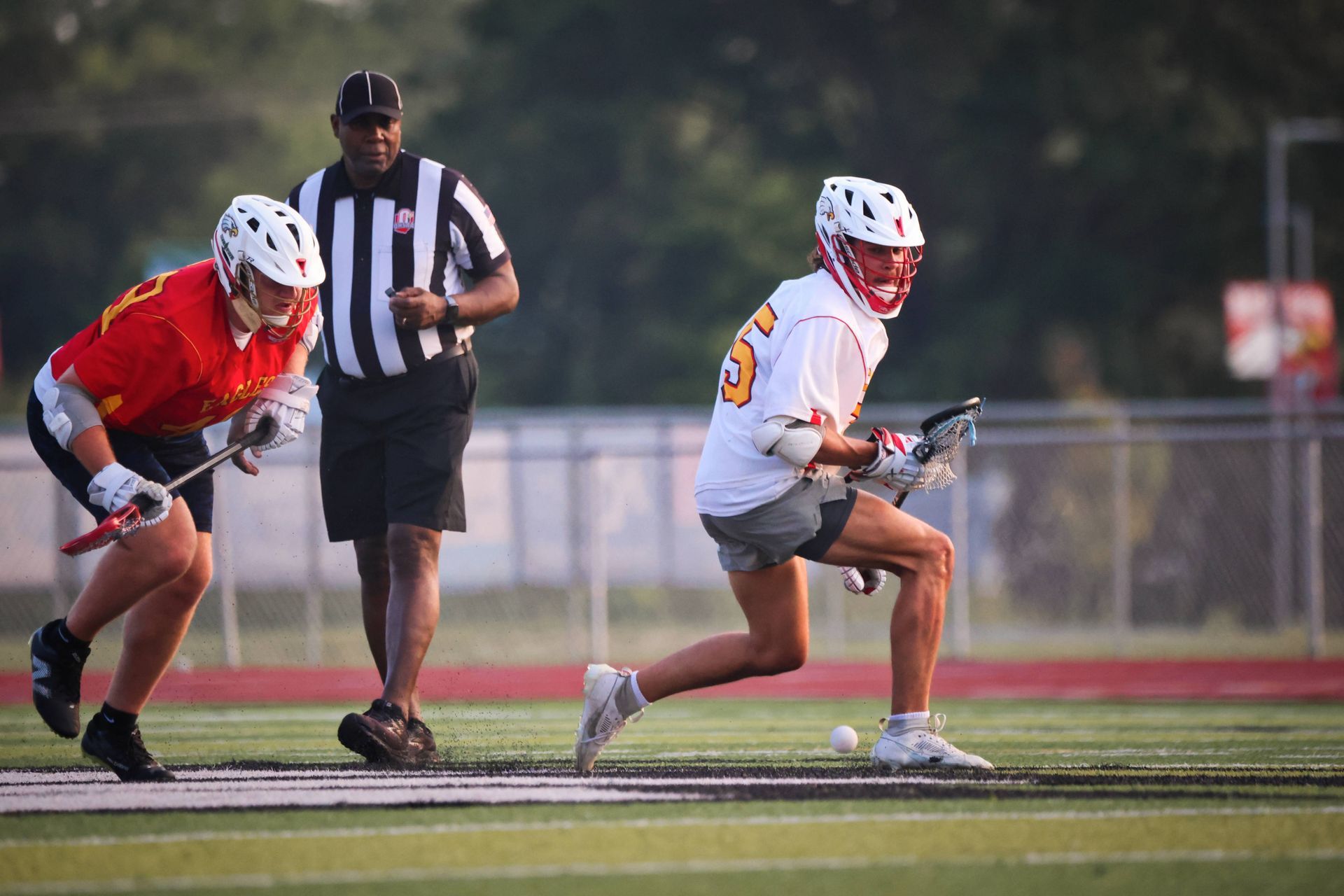 Two lacrosse players sprint on a field as a referee watches from behind