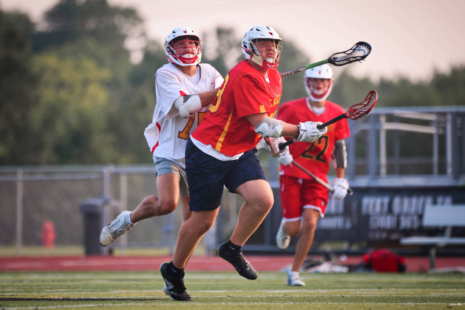 Lacrosse players in red and white sprint on a field, with one carrying the ball in the foreground.
