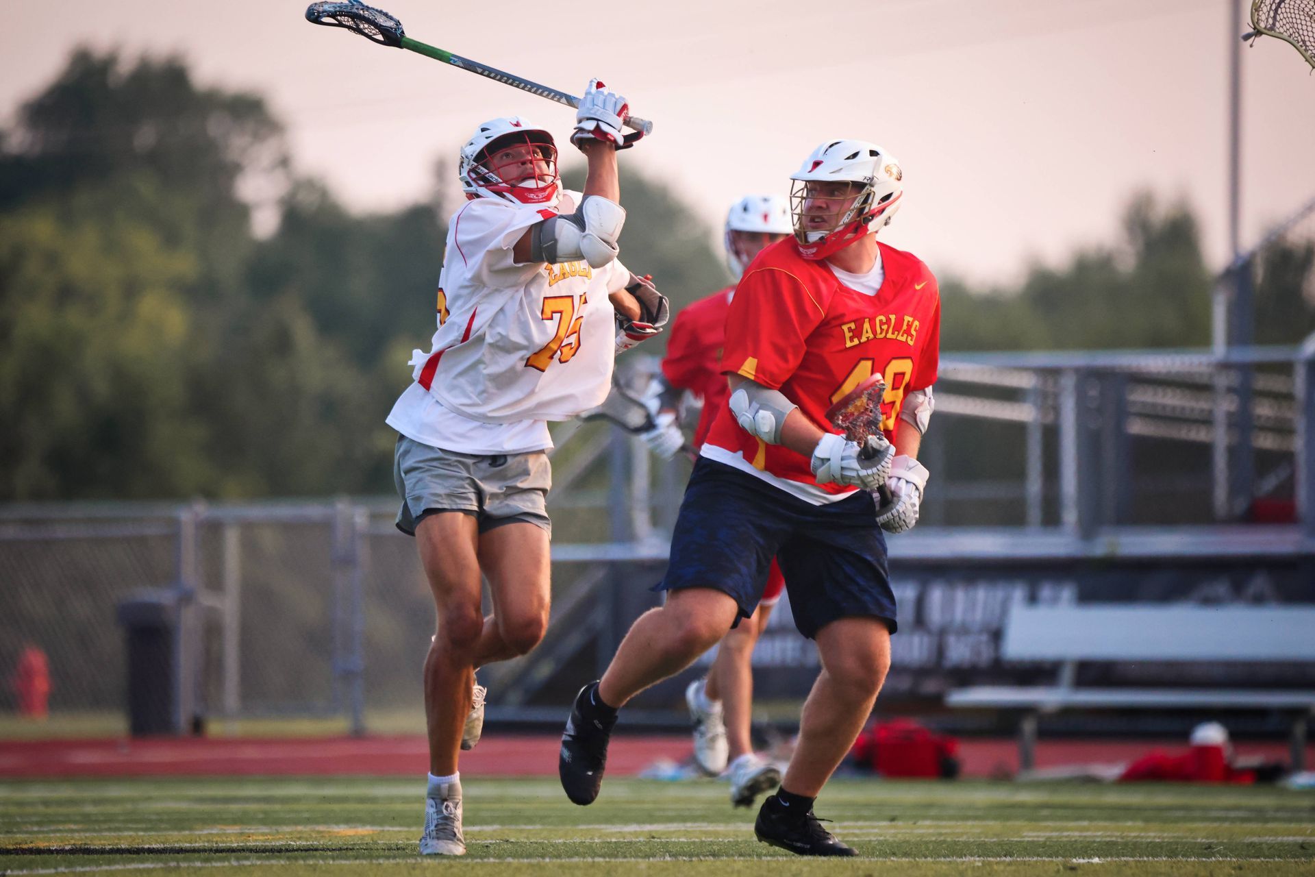 Two lacrosse players sprint on a field, one in white raising a stick over a defender in red.