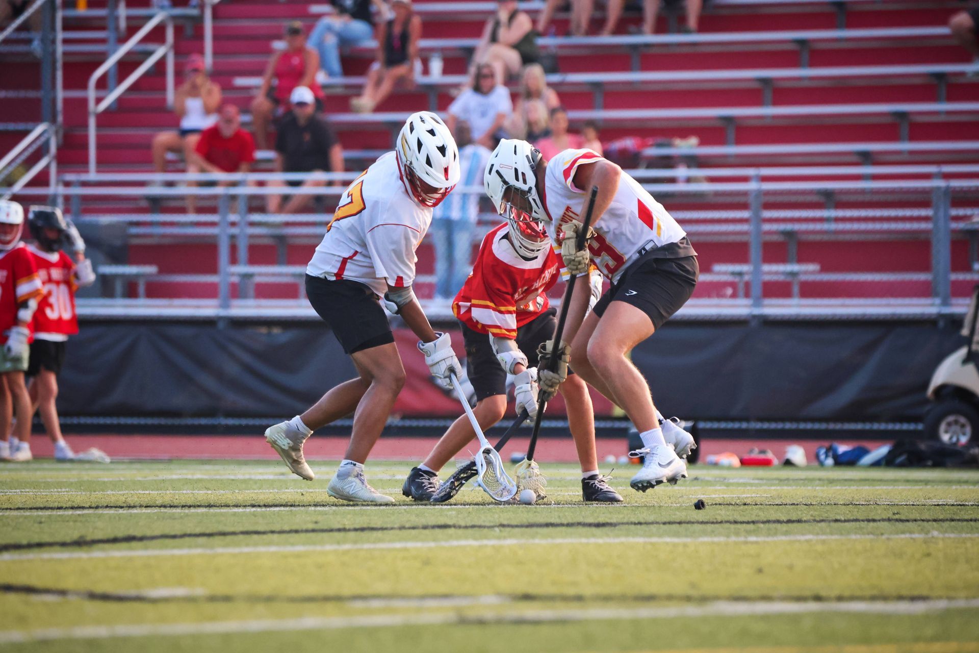 Three lacrosse players in white and red uniforms battle for the ball on a field