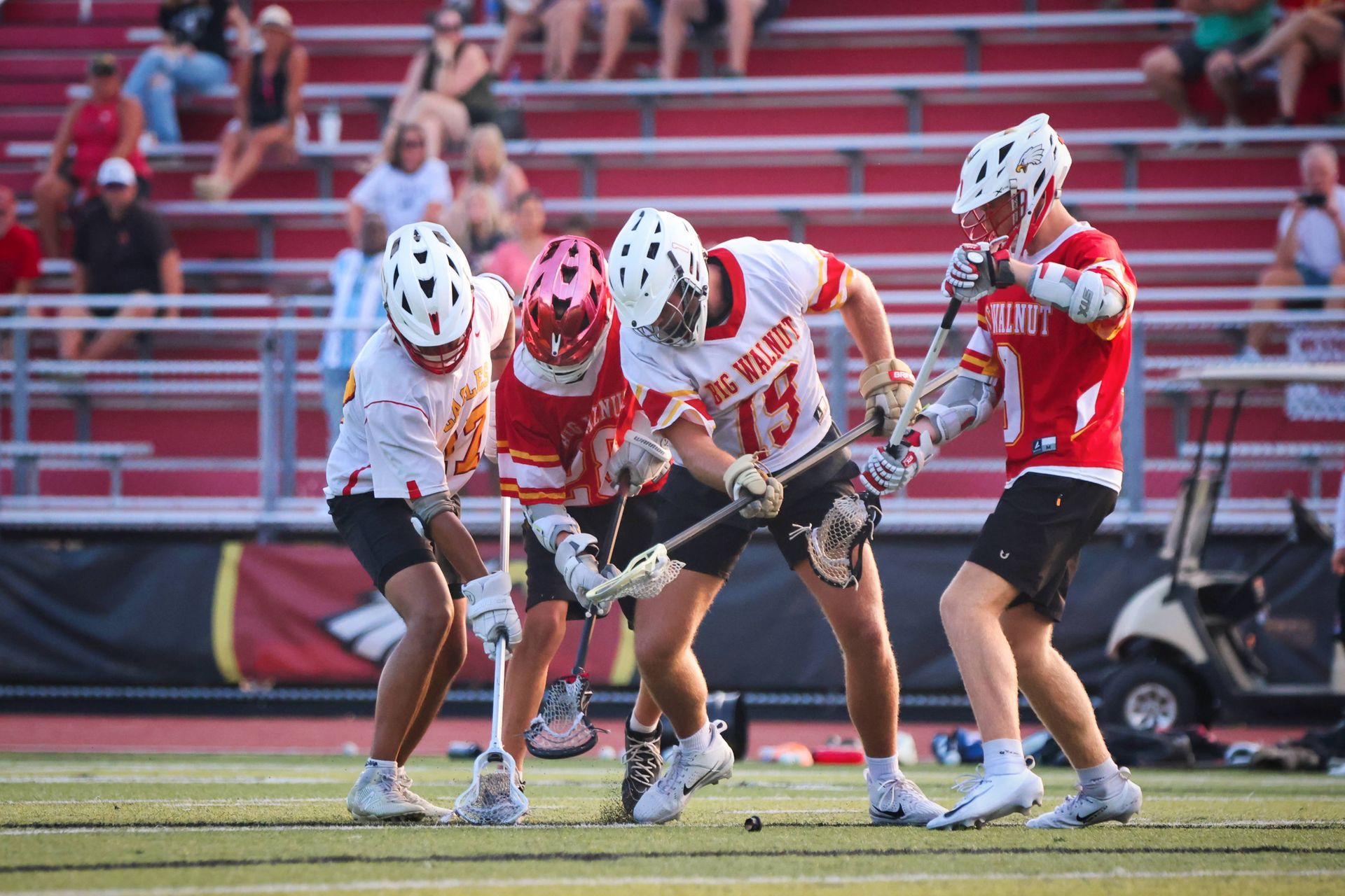 Lacrosse players in red and white uniforms battle for the ball on a field.