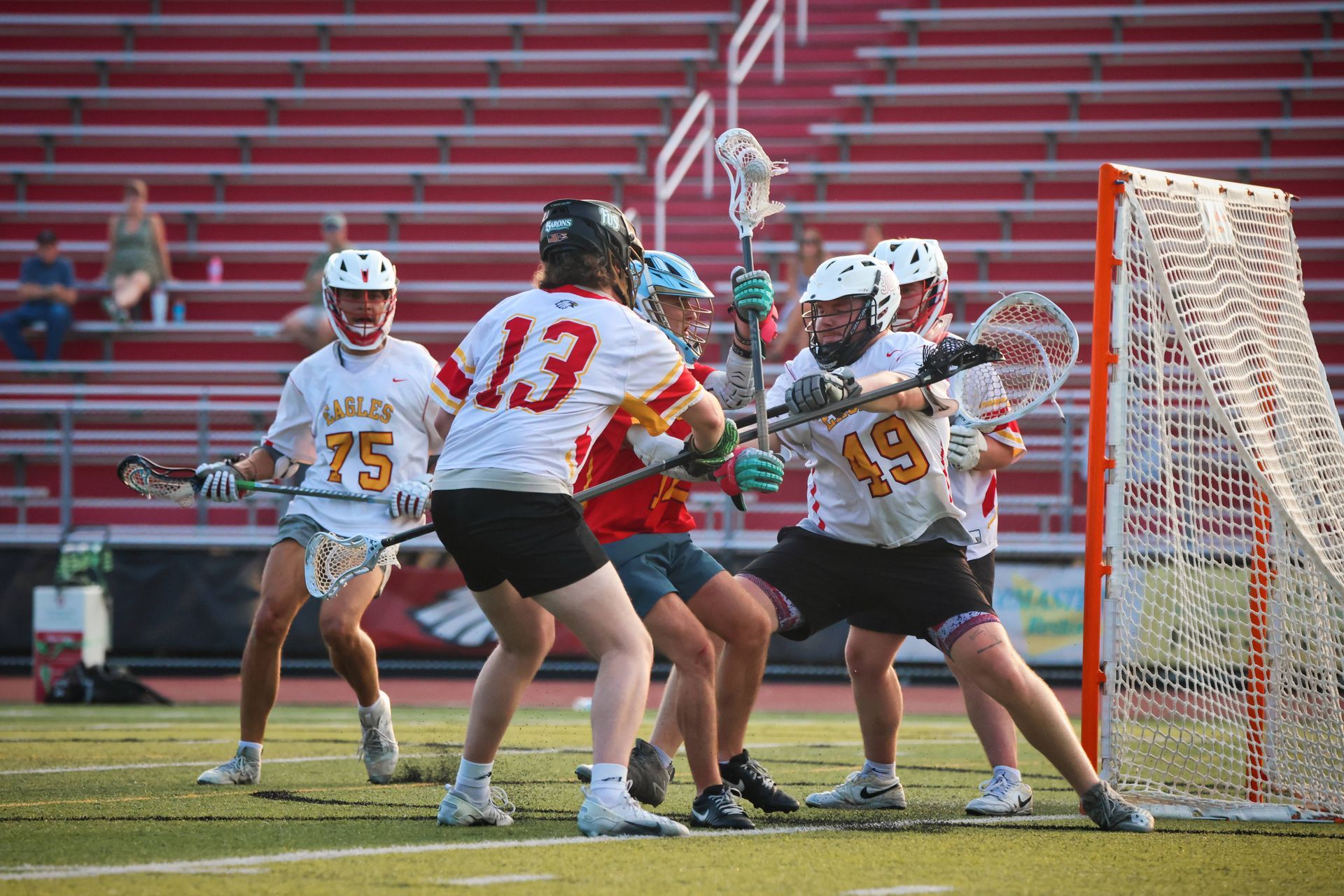 Lacrosse players in white and red jerseys battle near the goal on a field with bleachers behind them.