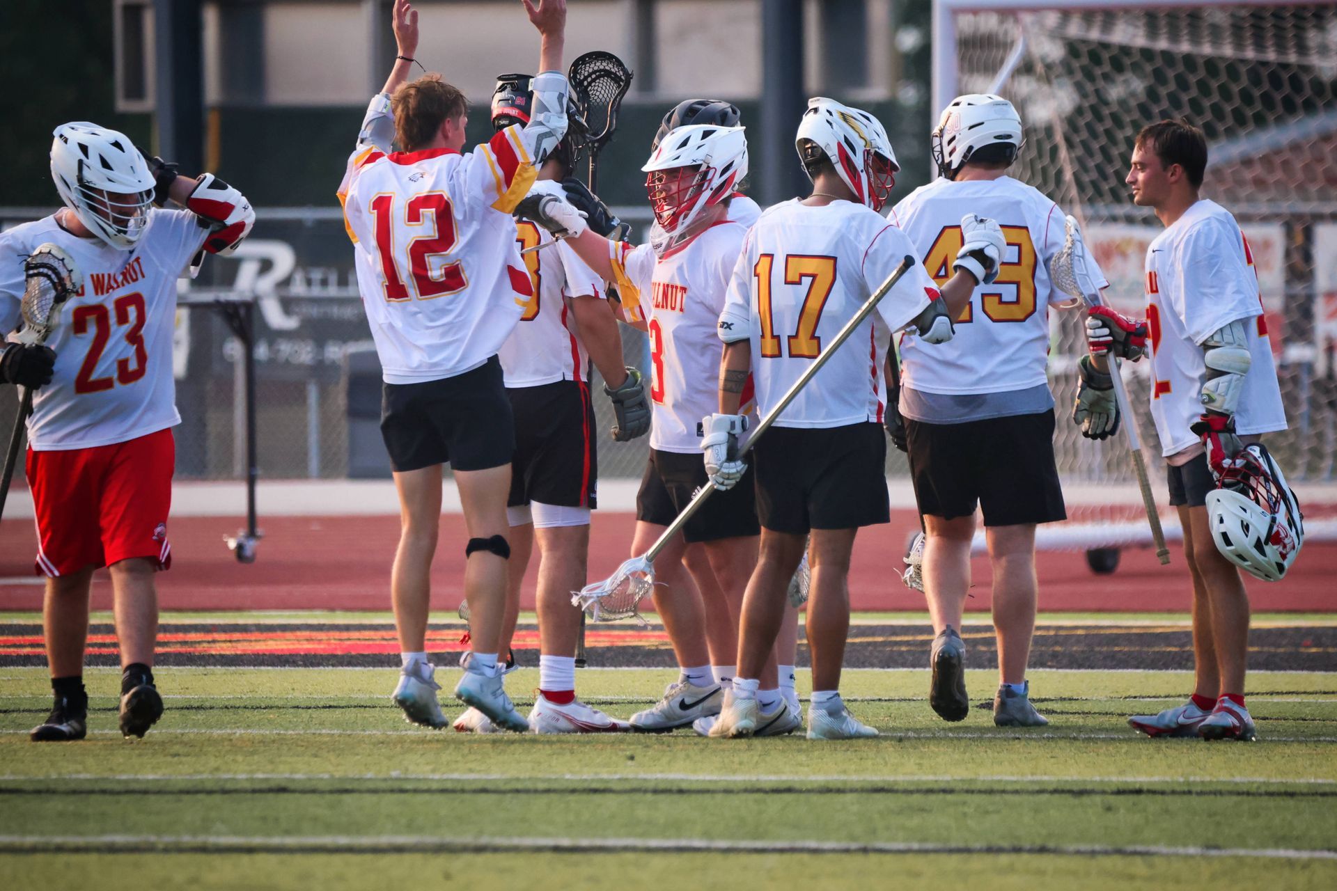 Lacrosse players in white and red jerseys huddle on a field after a play, one raising a hand.