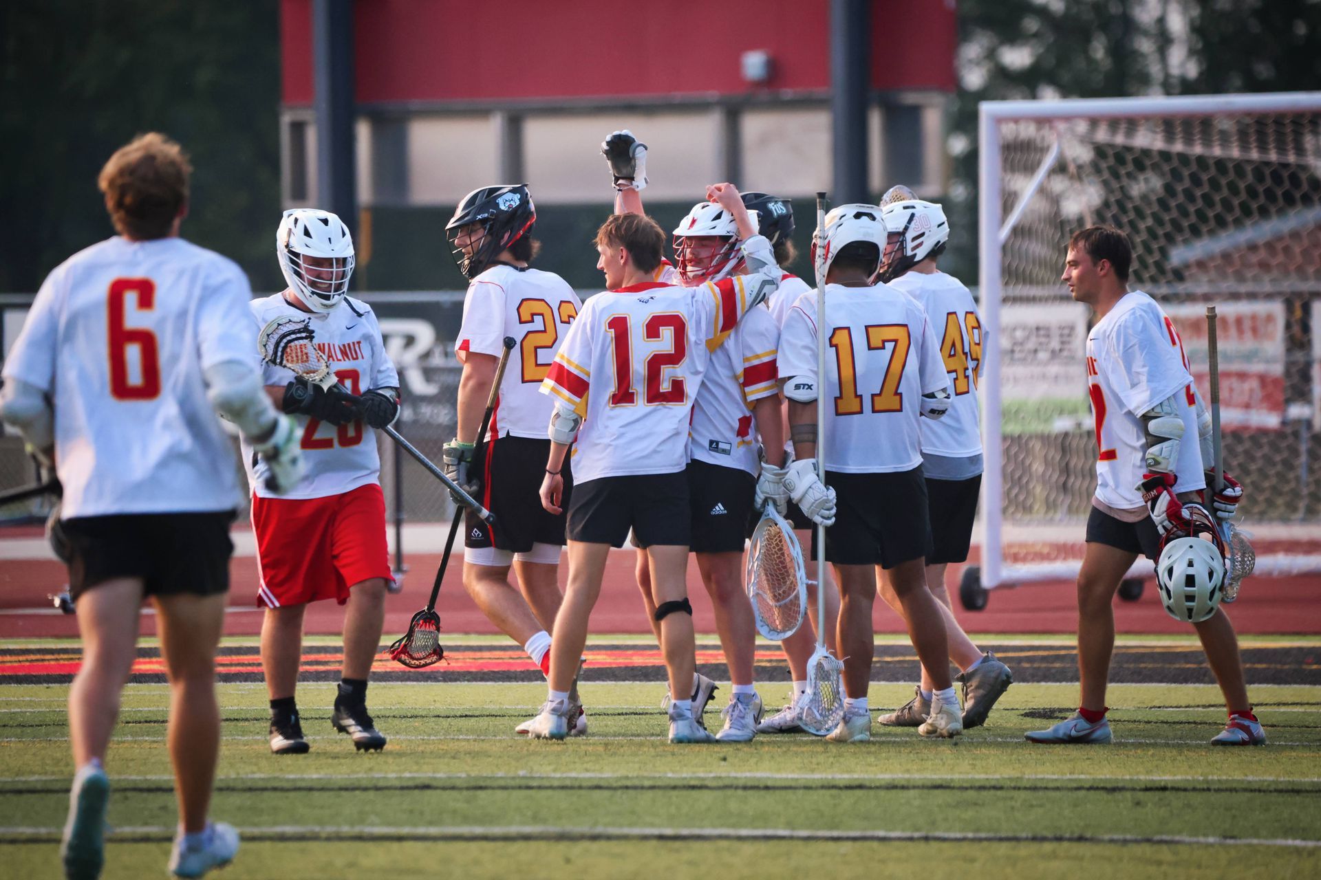 Lacrosse players in white jerseys huddle on a field near the goal, with one player raising a hand.
