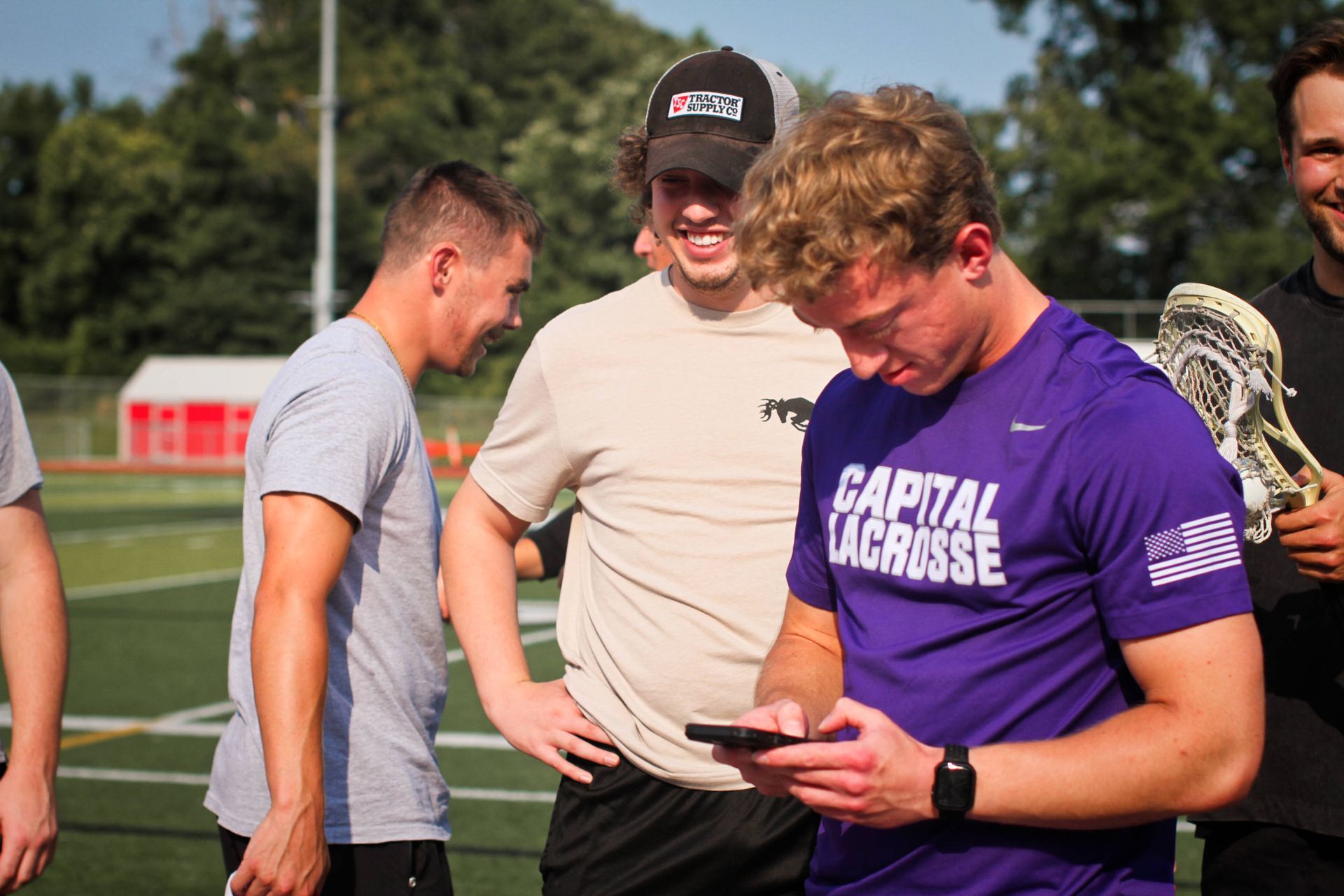 Three lacrosse players on a field, one in a purple Capital Lacrosse shirt holding a stick and looking at a phone.