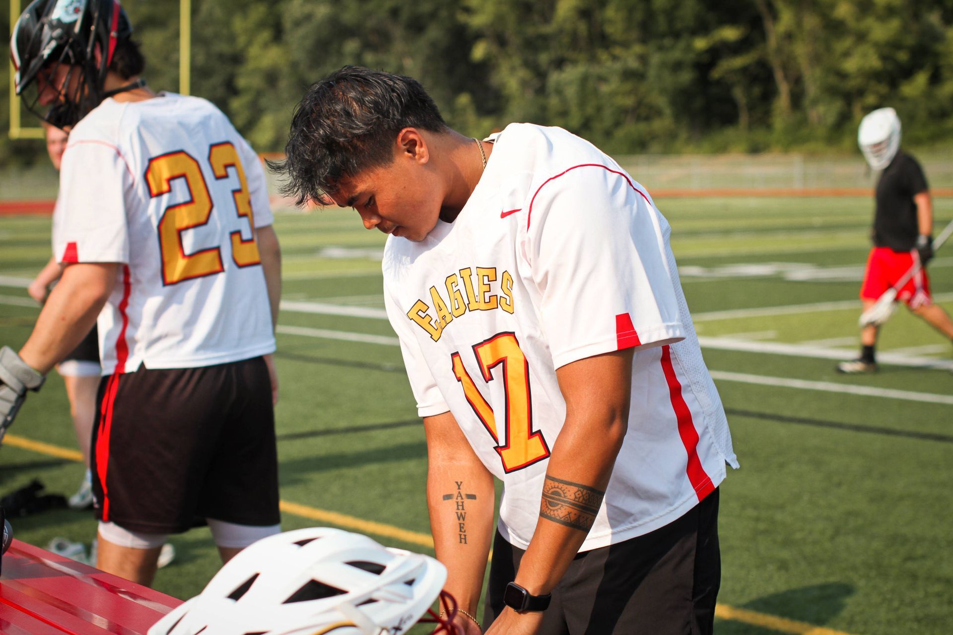 Two lacrosse players in white jerseys on a field, one bent over adjusting gear, helmets and net visible