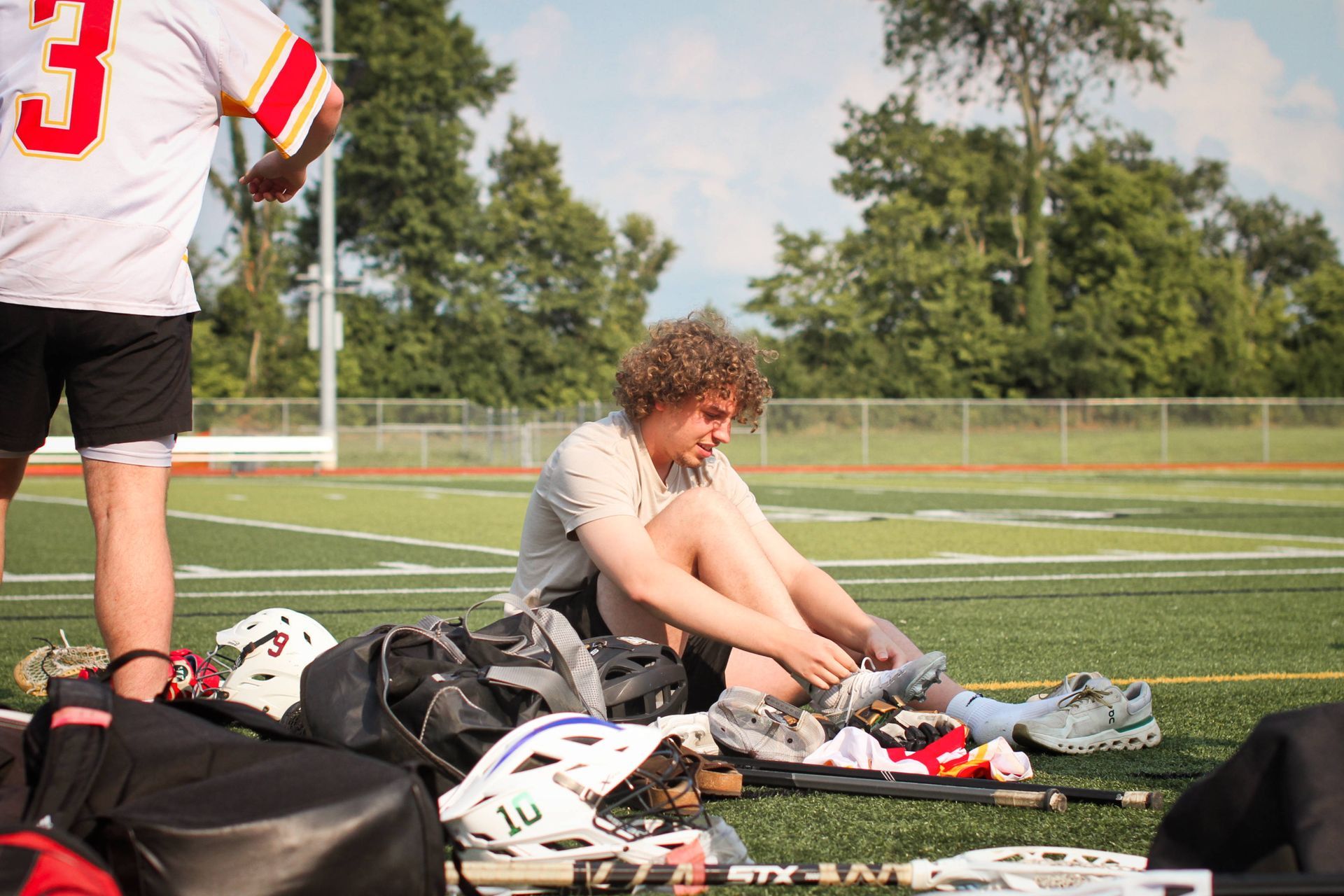 Lacrosse player sitting on the field tying cleats beside a pile of gear during practice