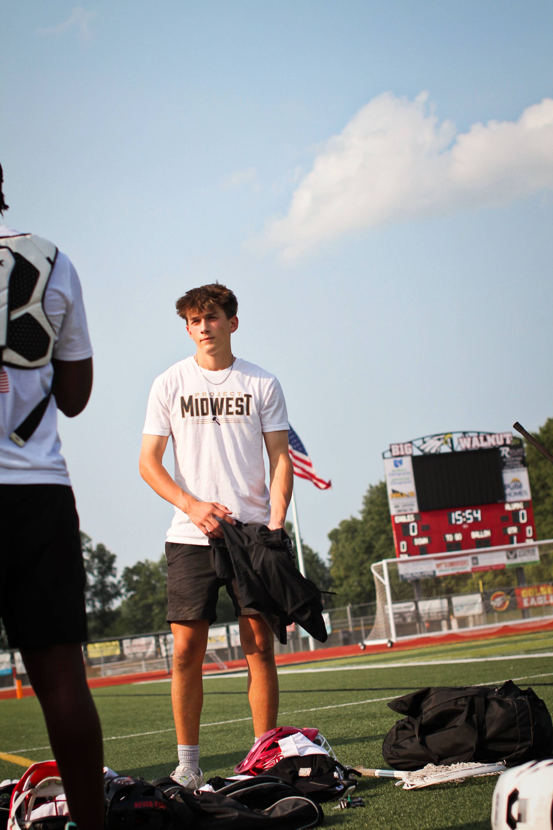 Teen in white T-shirt and black shorts stands on a track field beside sports gear, under a blue sky.