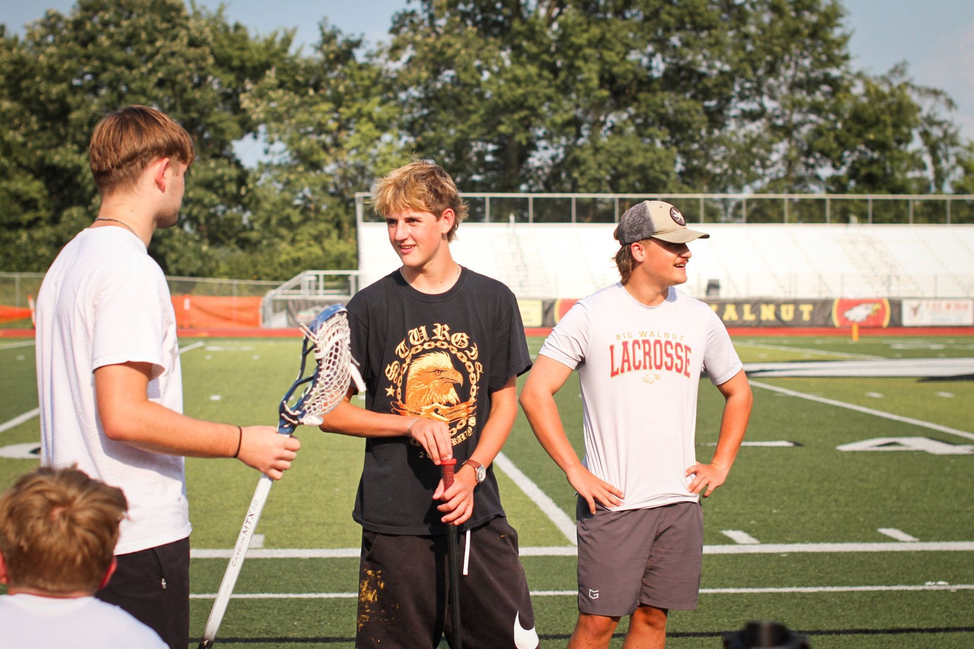 Three people stand on a sports field, two holding lacrosse sticks, as they talk after a game.