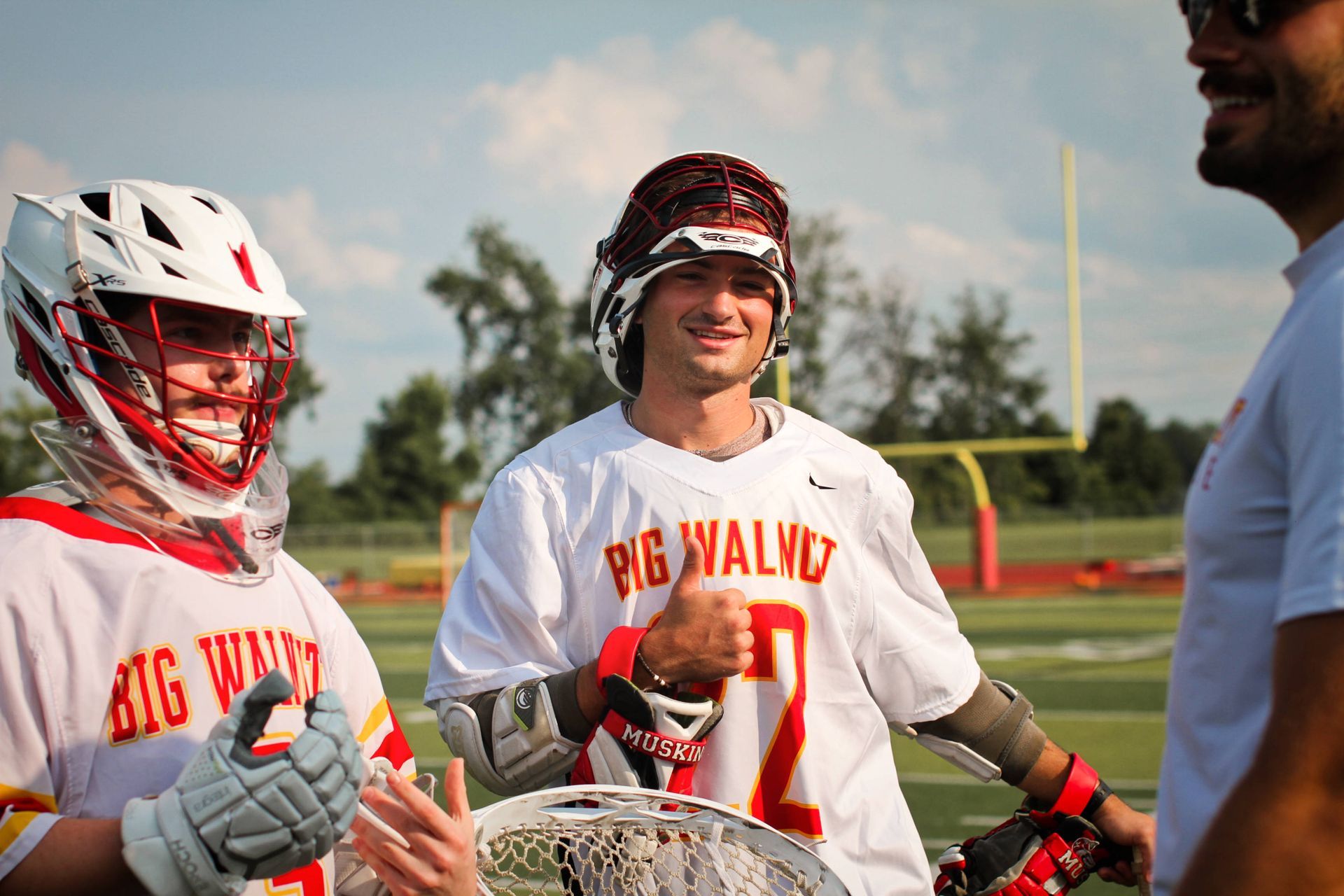 Two lacrosse players in white uniforms and helmets stand on a field, talking and smiling with a coach nearby.