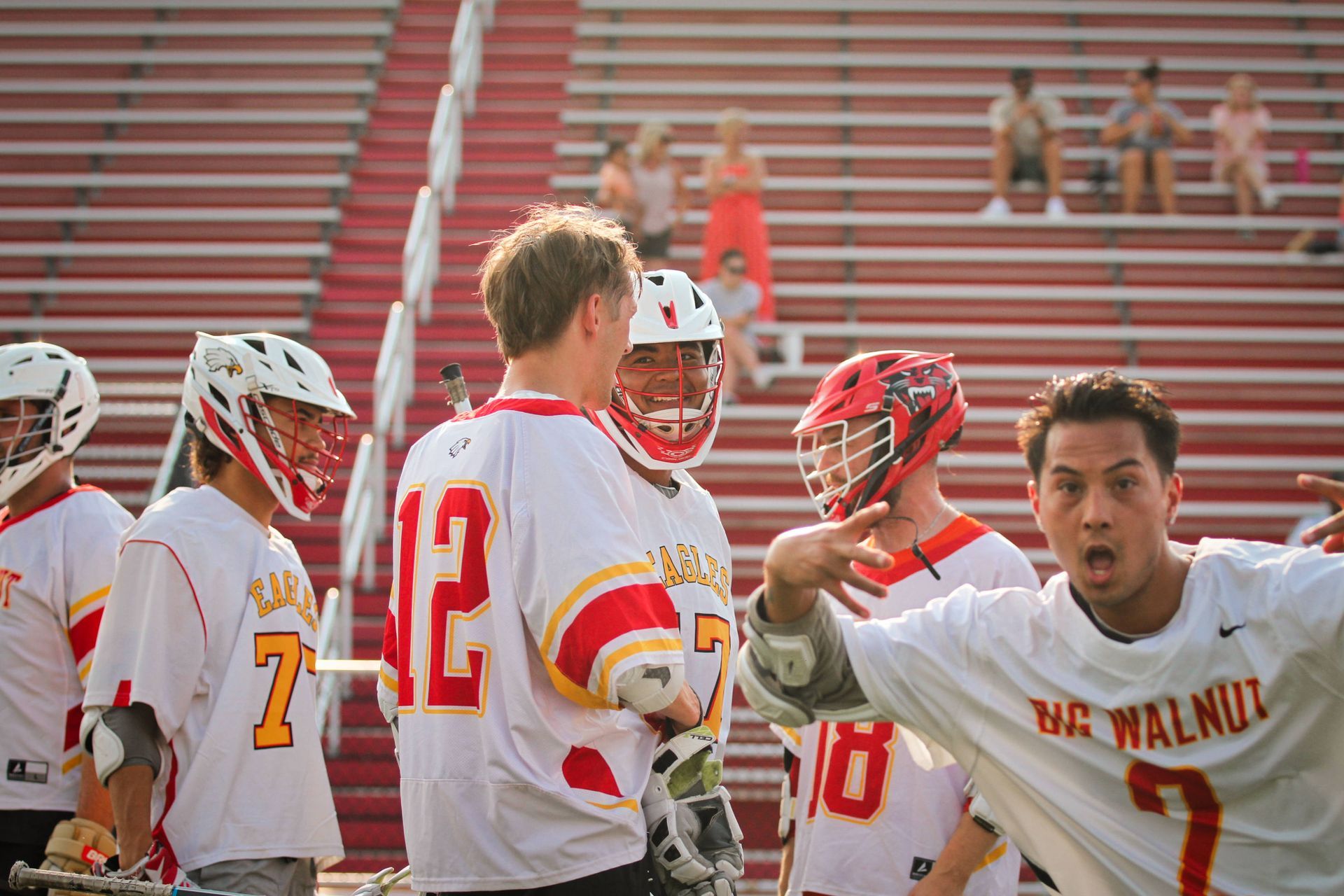 White-and-red lacrosse players huddle on the field, one shouting with a raised hand in front of stadium bleachers.