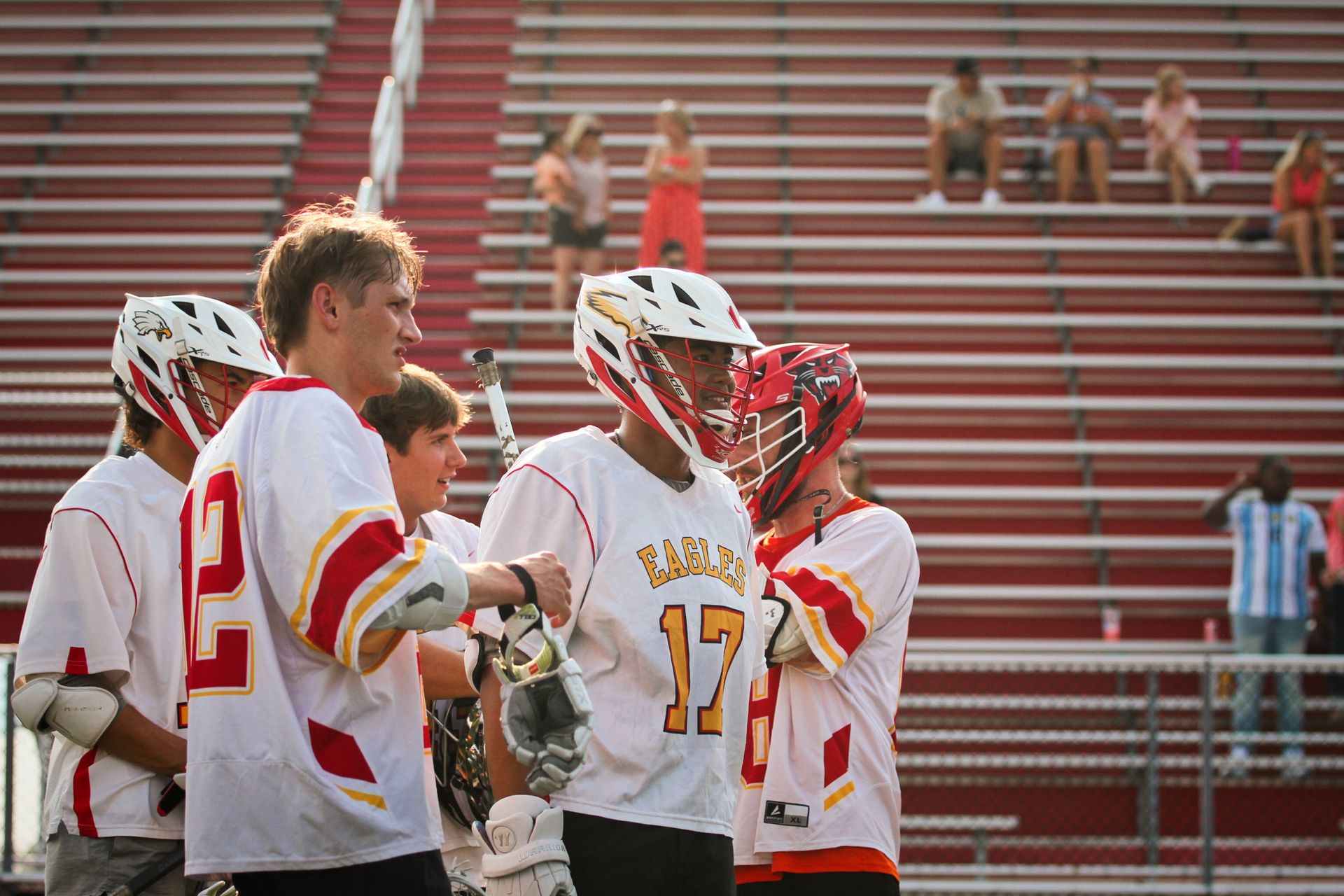 Lacrosse players in white jerseys with red helmets huddle on the field, with empty bleachers behind them