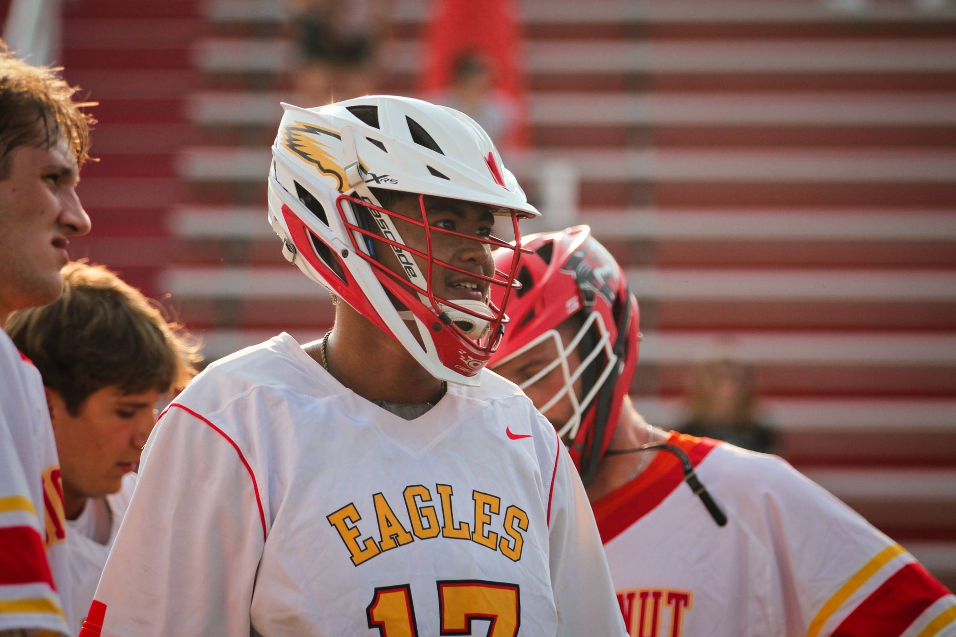 Lacrosse player in white Eagles jersey and helmet, standing on a field with teammates in the background
