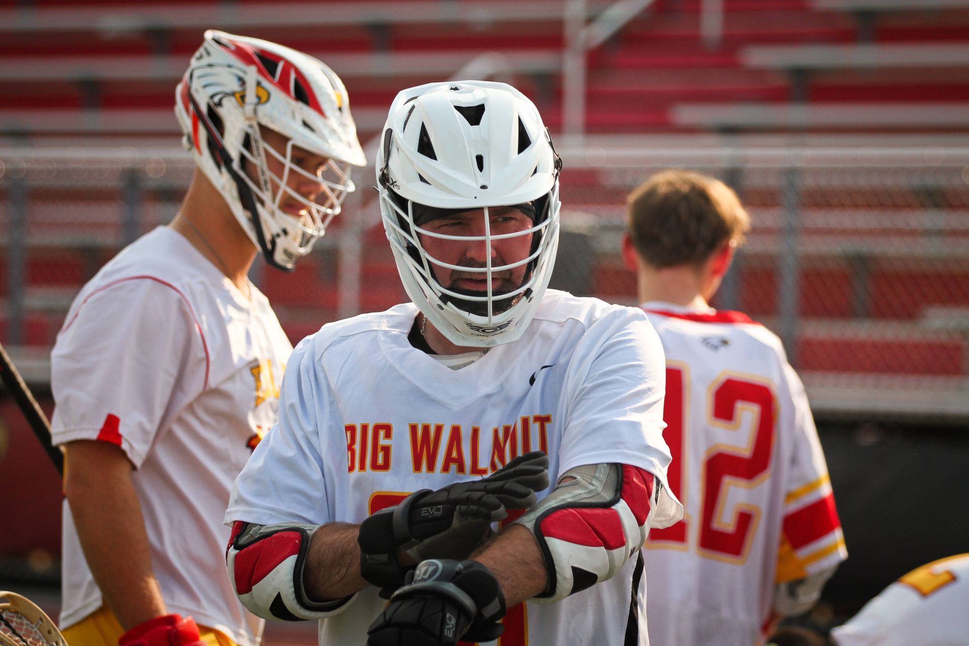 Lacrosse players in white and red uniforms standing on a field; one looks down at his gloves.