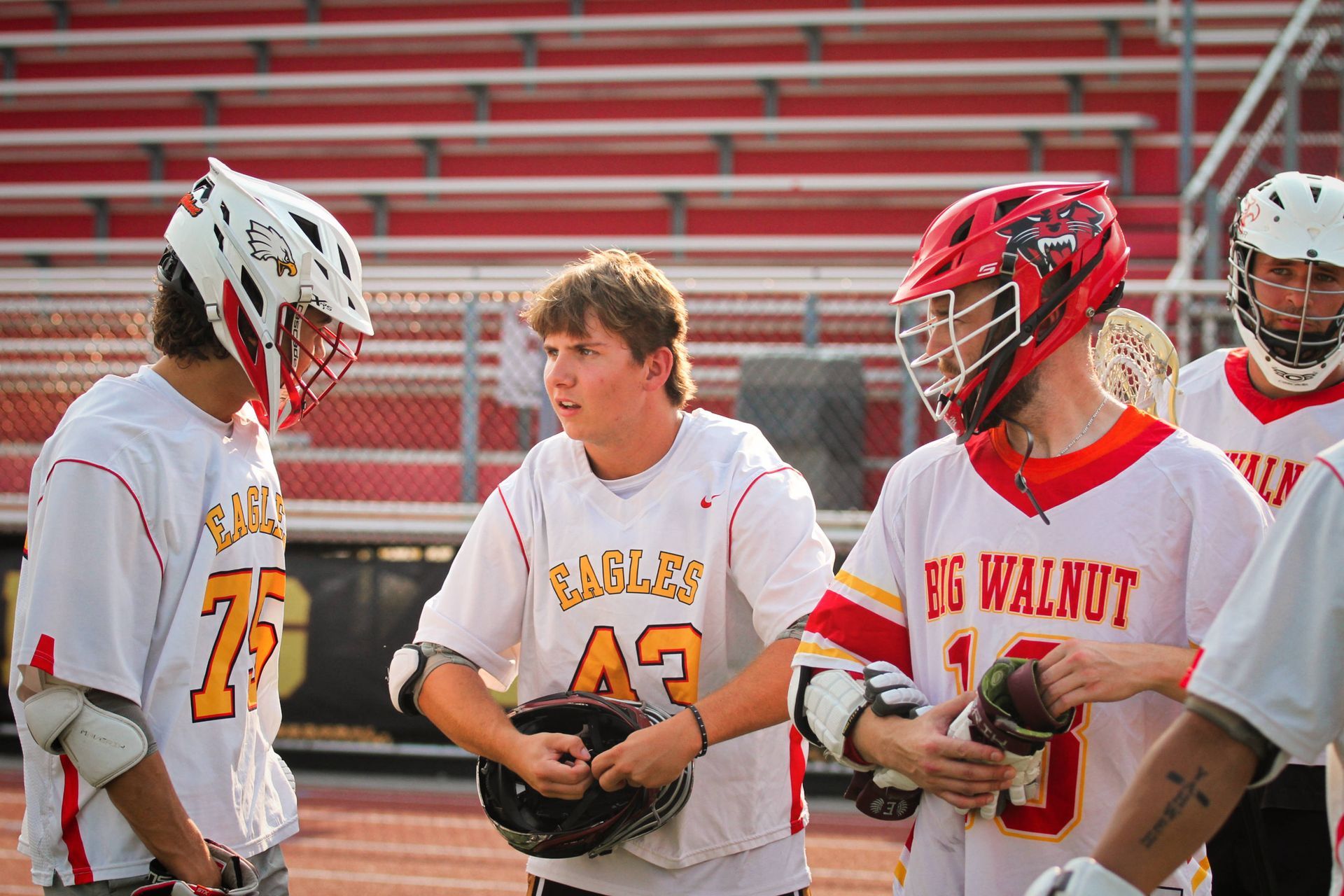 Former high school lacrosse players in white and red helmets stand on the field during a game timeout