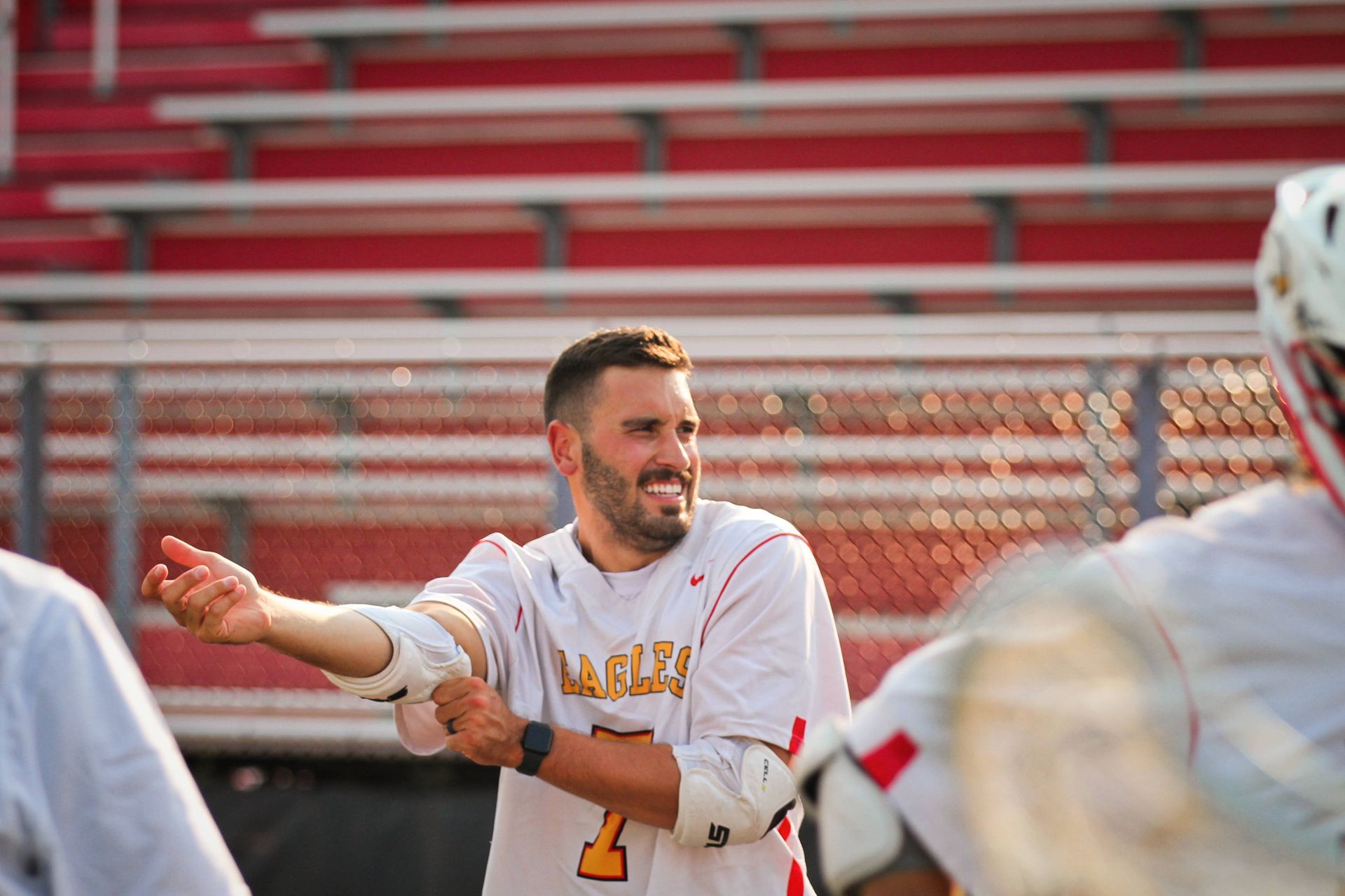 Lacrosse player in white jersey pointing and smiling on the field, with teammates blurred in the foreground