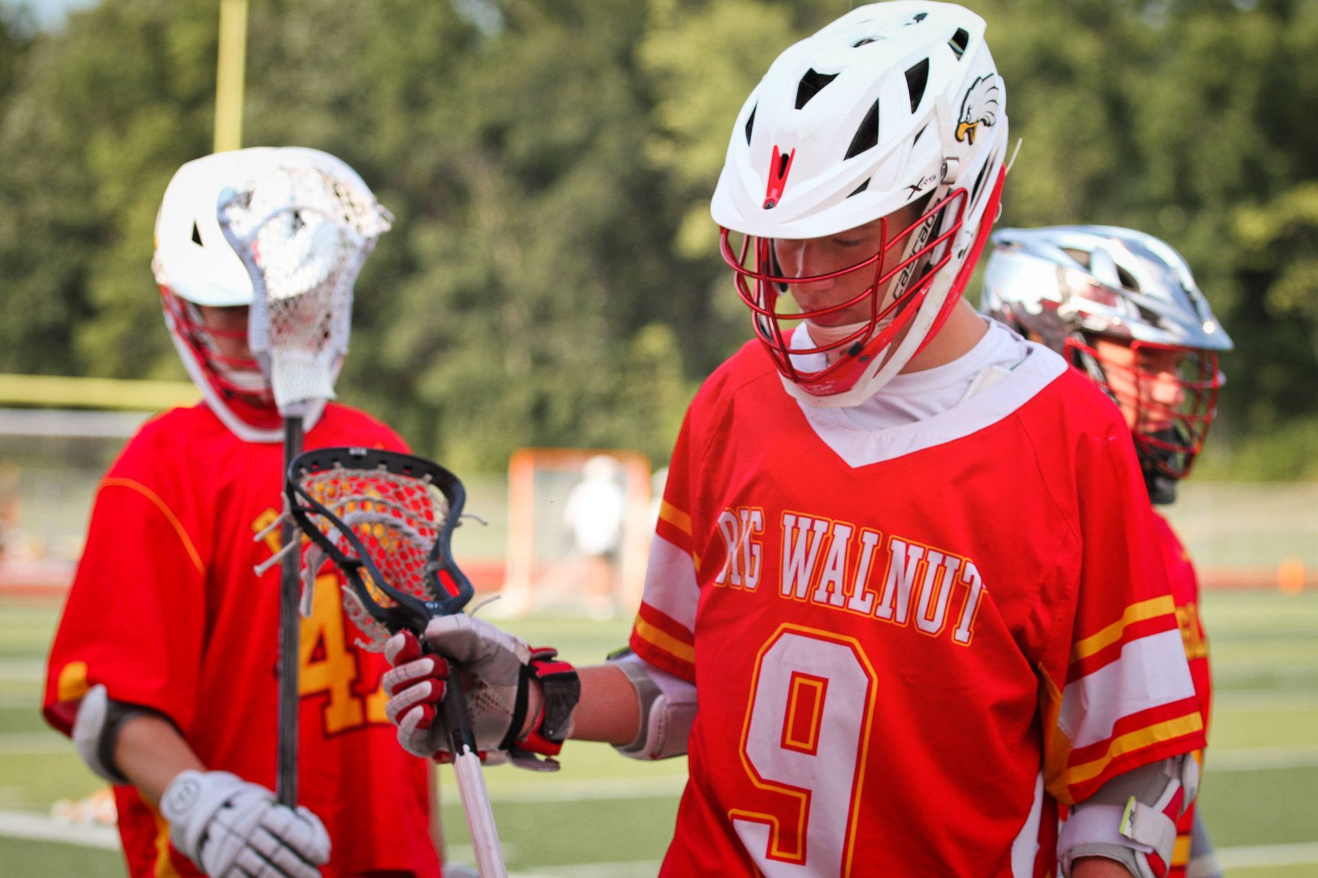 Lacrosse players in red jerseys and white helmets standing on a field, one checking a stick.