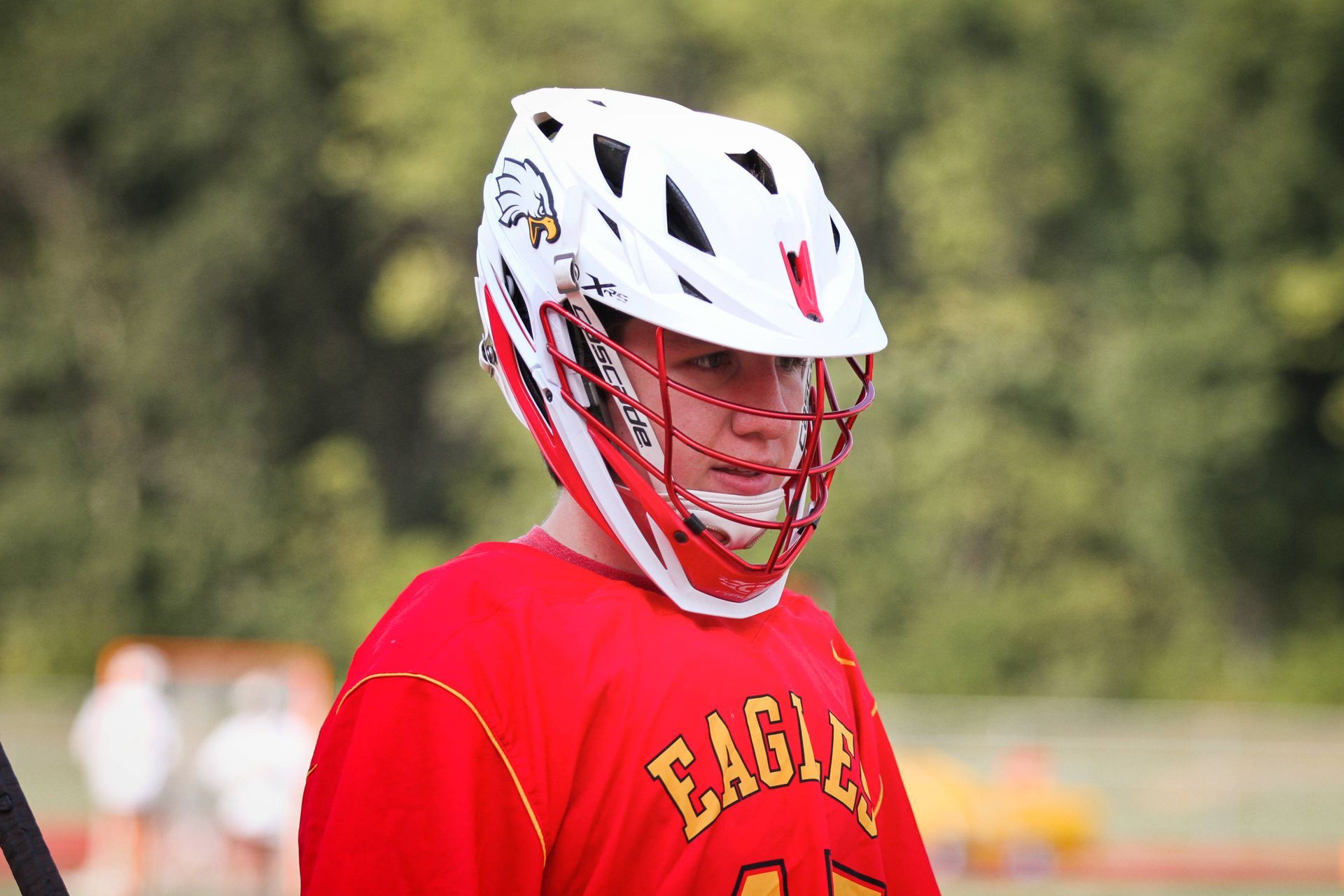 Lacrosse player in red jersey and white helmet, looking down on a field
