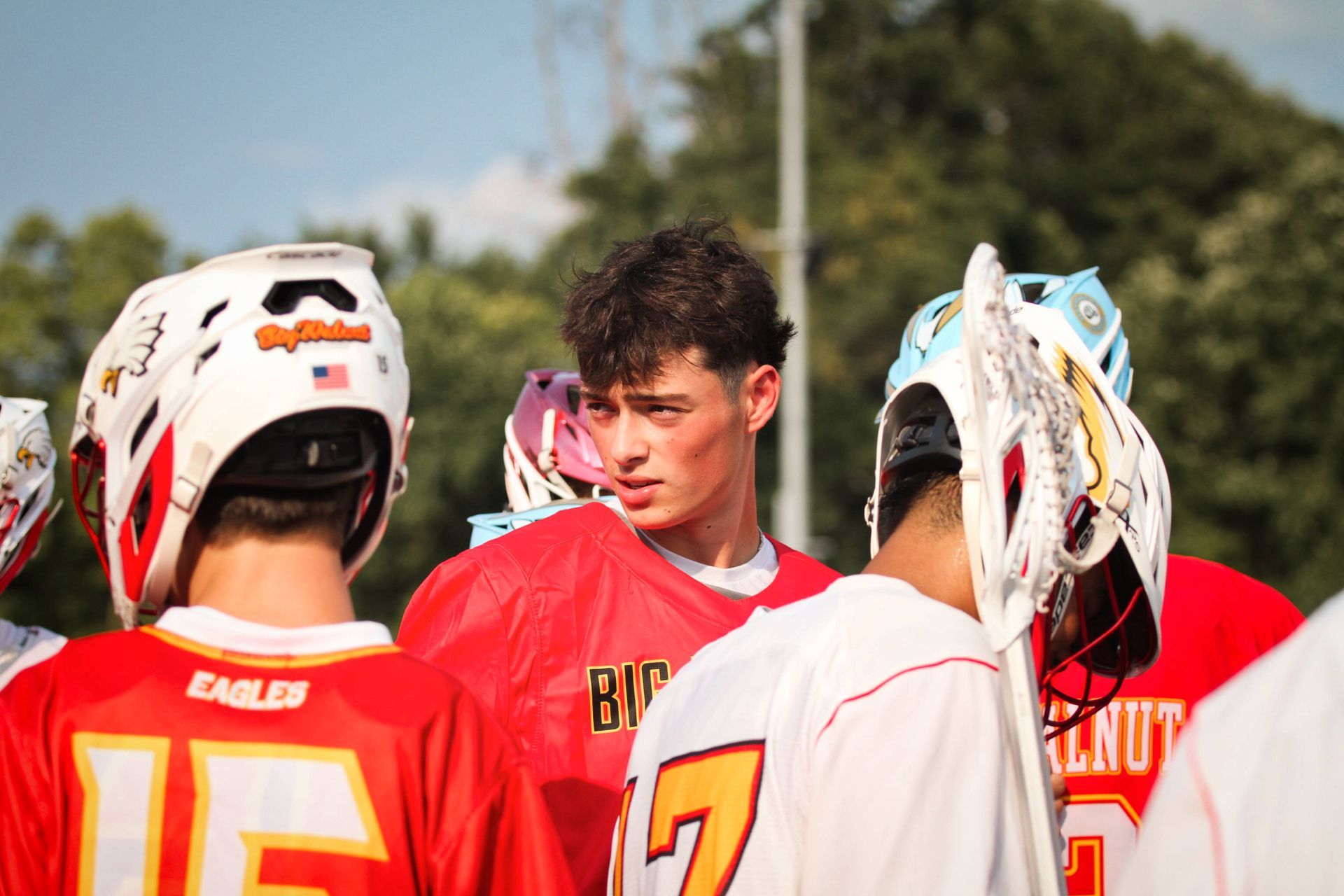 Lacrosse players in red and white jerseys huddle on a field, listening to a teammate.