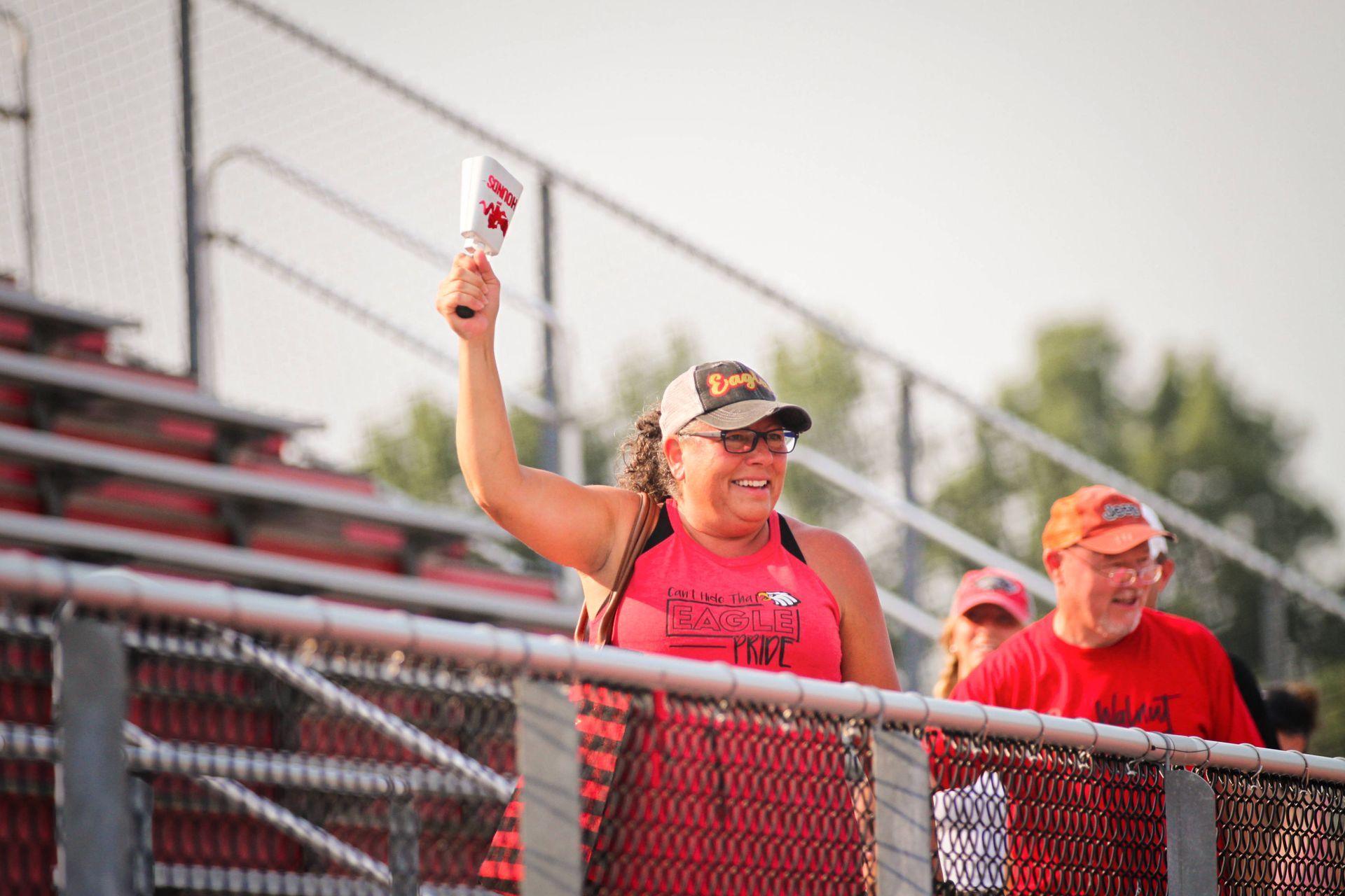 Two fans in red shirts cheer in the bleachers, one holding a cowbell aloft behind a chain-link fence.