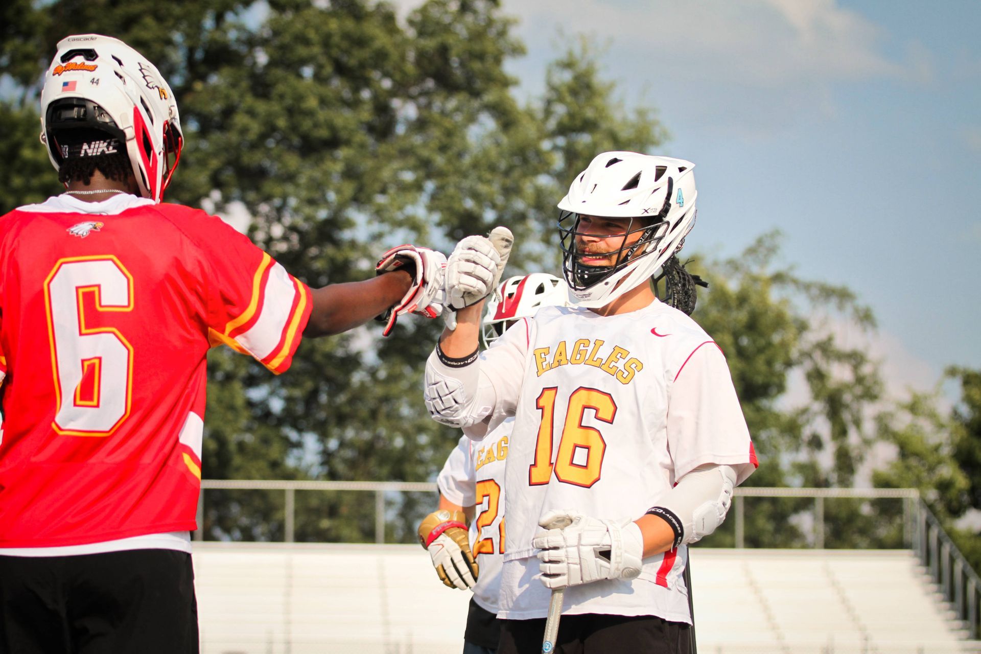 Two lacrosse players in white and red jerseys fist-bump on a field under a bright sky.