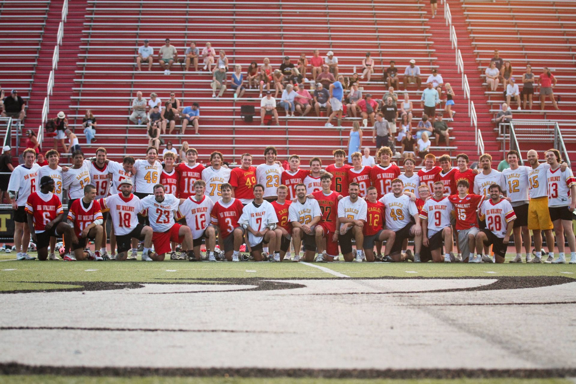 Lacrosse team posing on a field in red-and-white uniforms with stadium bleachers behind them