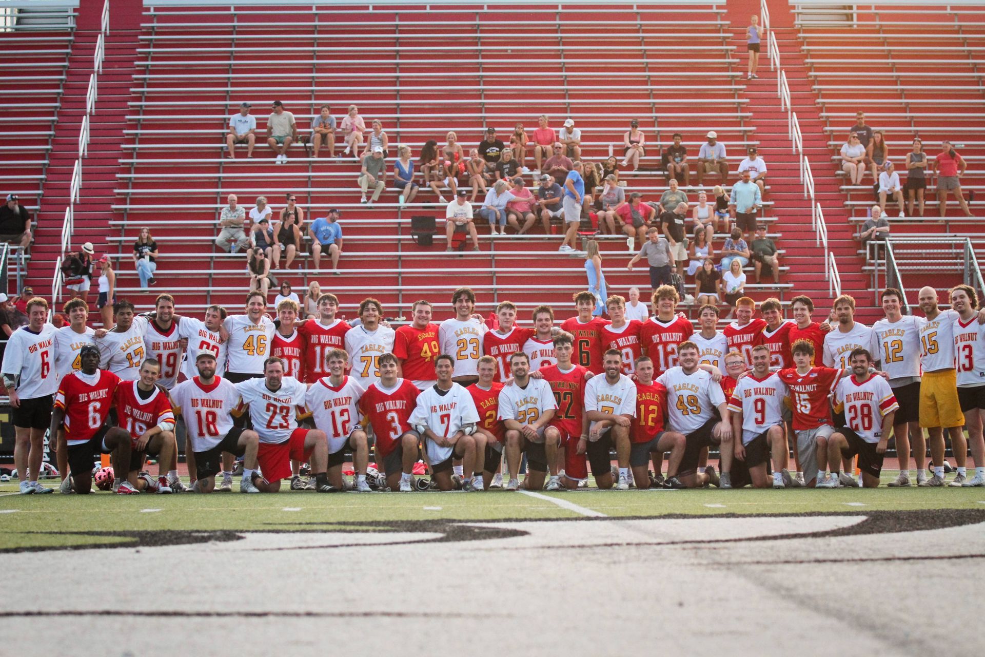 Lacrosse team posing on a stadium field in red and white uniforms, with fans in the bleachers behind them
