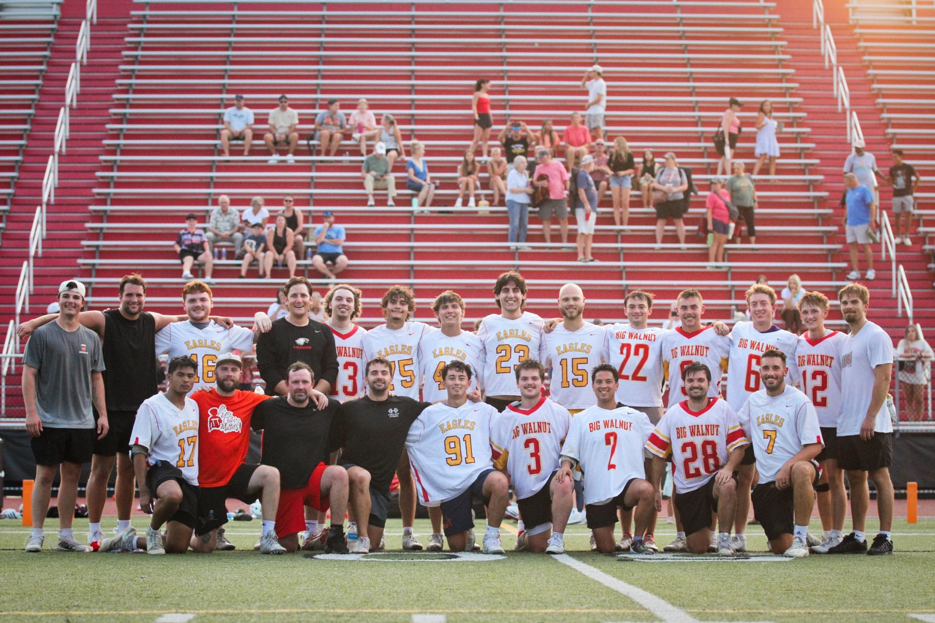 Lacrosse team posing on a field in white and red uniforms in front of stadium bleachers
