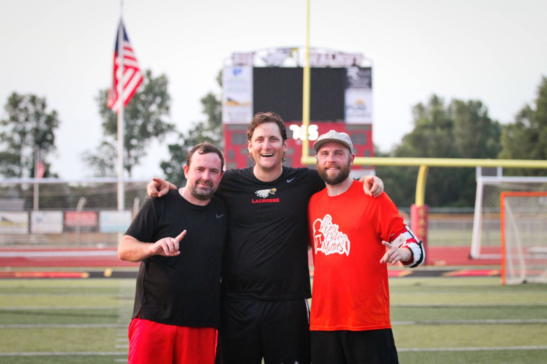 Three smiling people on a soccer field, standing arm-in-arm in front of the goal and scoreboard.