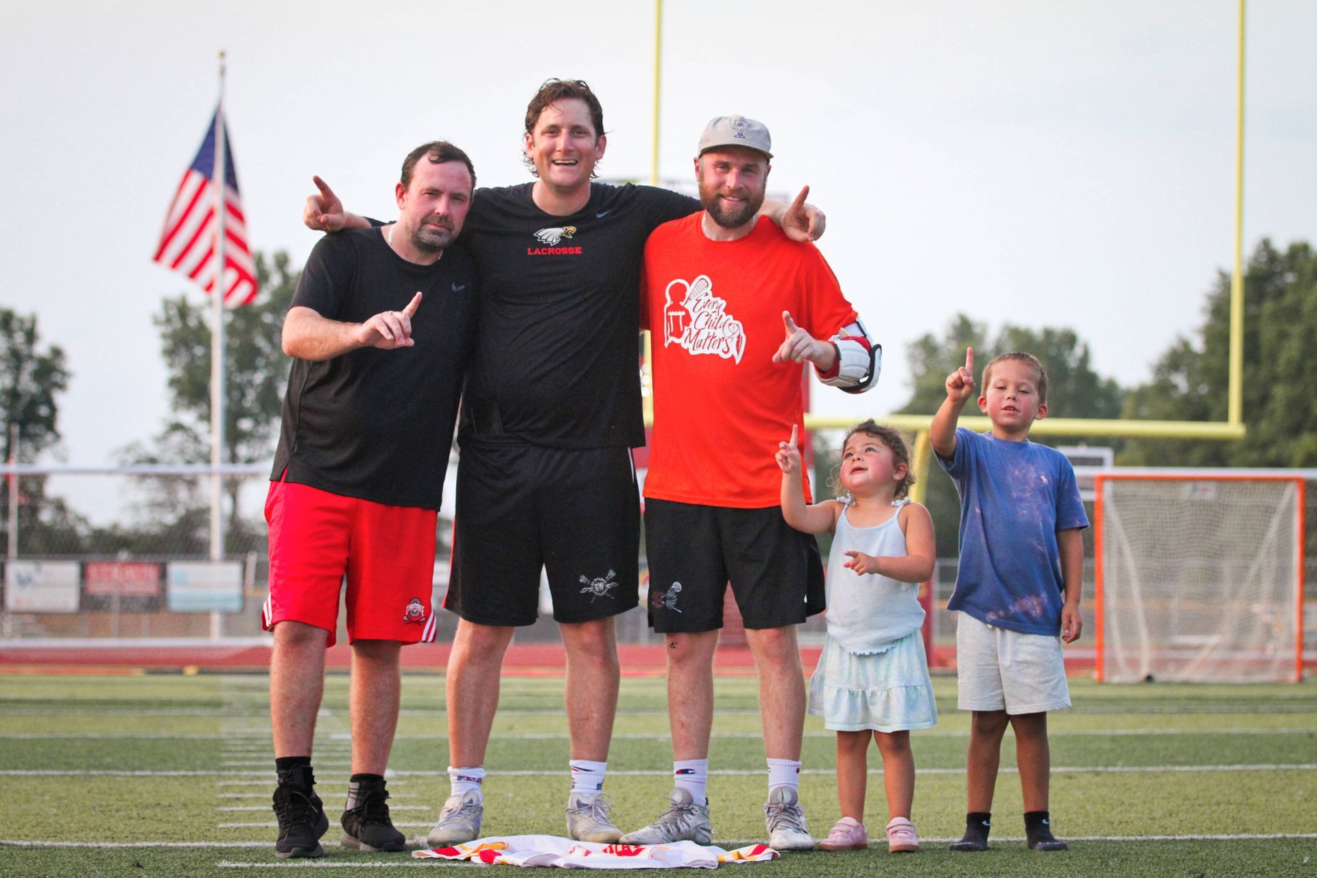 Four people pose on a lacrosse field, standing on a medal podium with an American flag in the background.