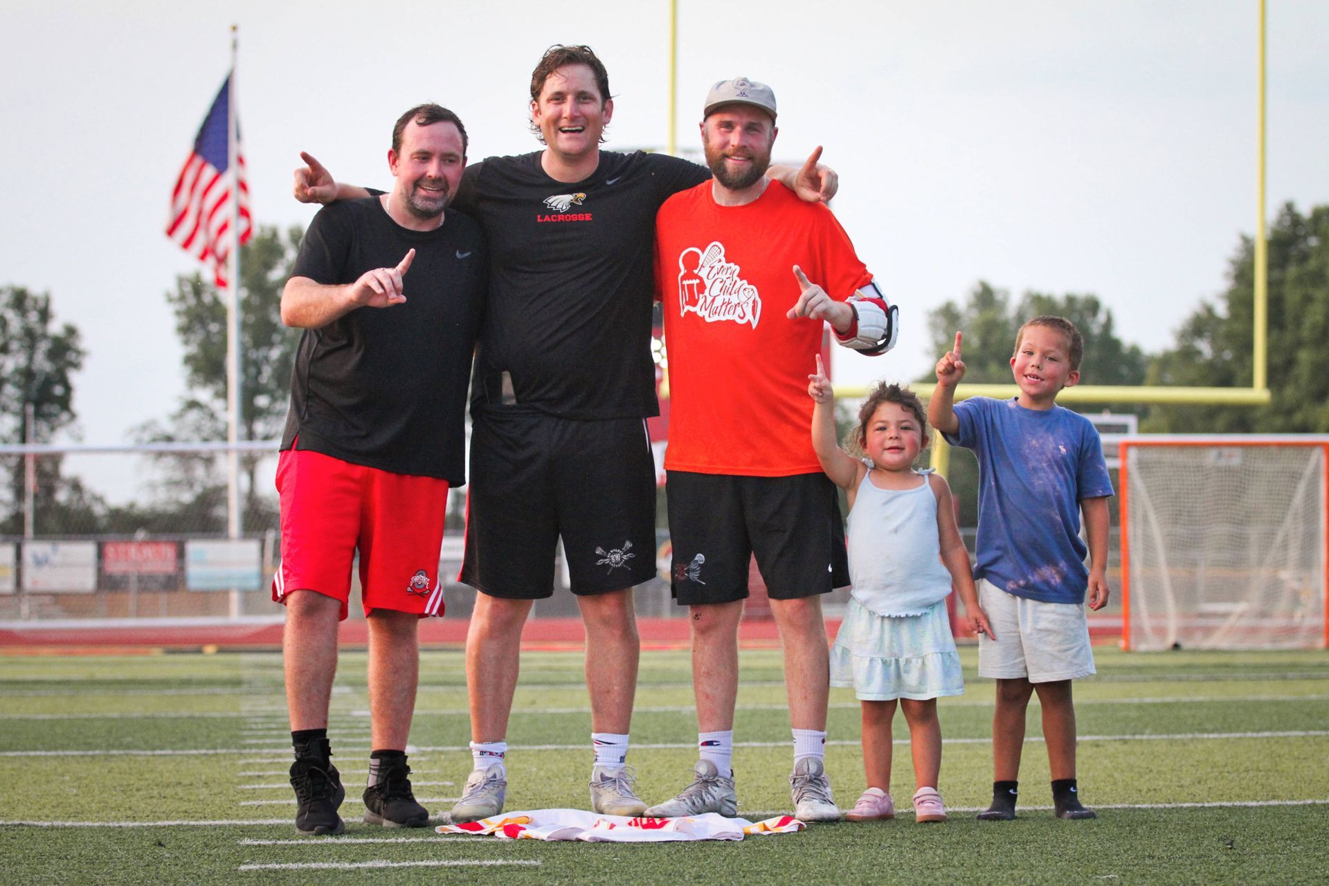 Three men pose on a football field with two children near a goal post and an American flag in the background.