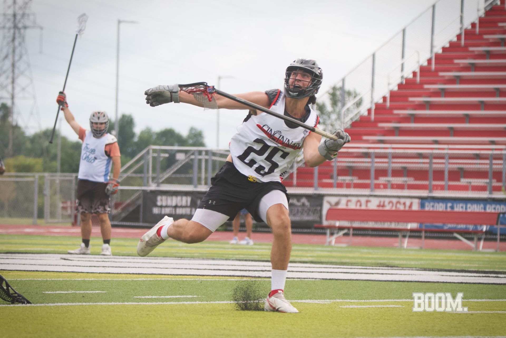 Lacrosse player in white and black jersey running on wet field, stadium seats in background