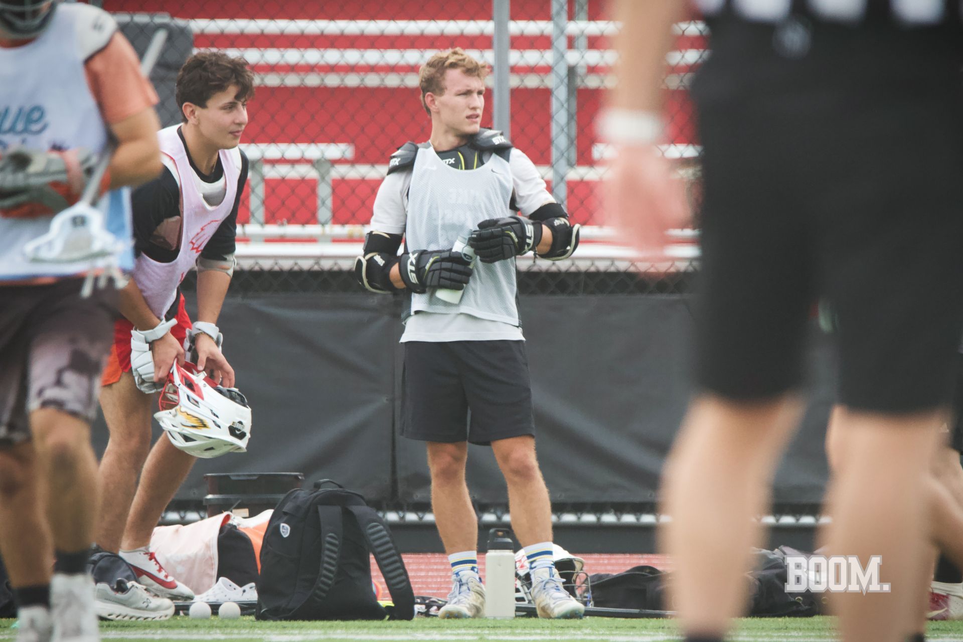 Lacrosse practice on a field, with players stretching and a coach holding papers near red stadium seats