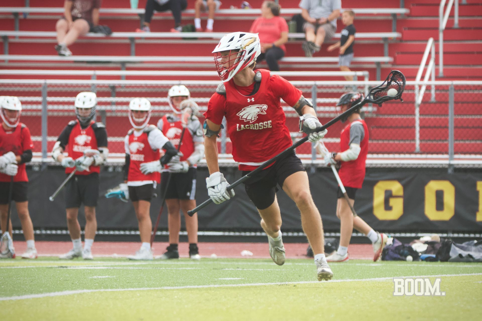 Lacrosse player in red sprints with the ball during a game, teammates and bleachers behind her.