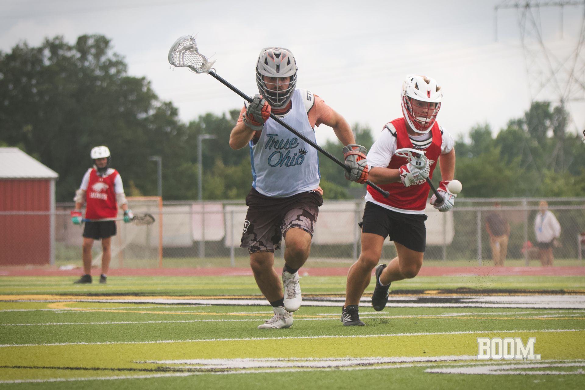 Lacrosse players sprinting on a wet field, one in gray chasing a player in red and white.