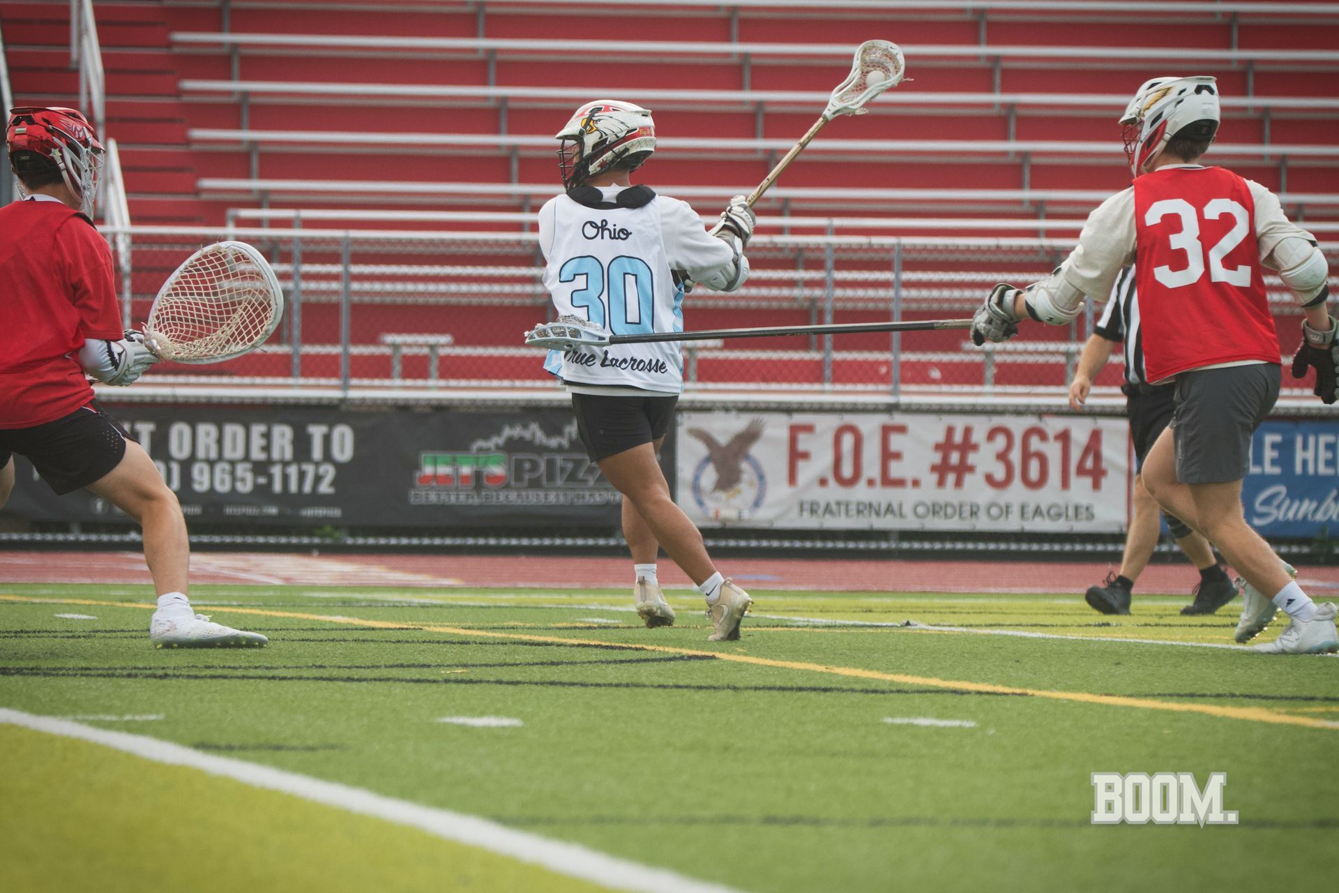 Lacrosse players in red and white sprint across the field during a game, with one player mid-shot near the goal.