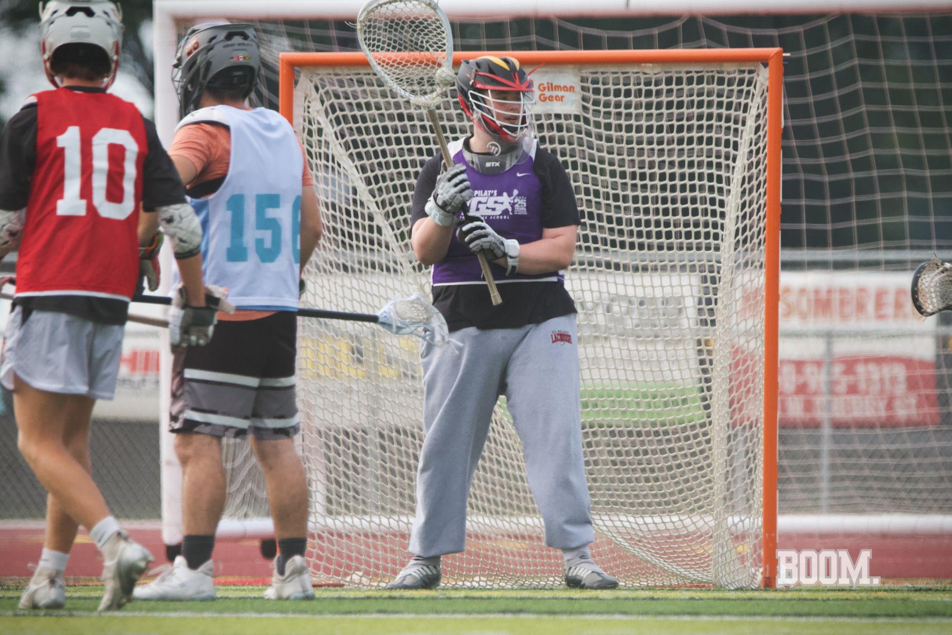 Lacrosse goalie in purple and gray stands in front of the net as two players face him nearby.