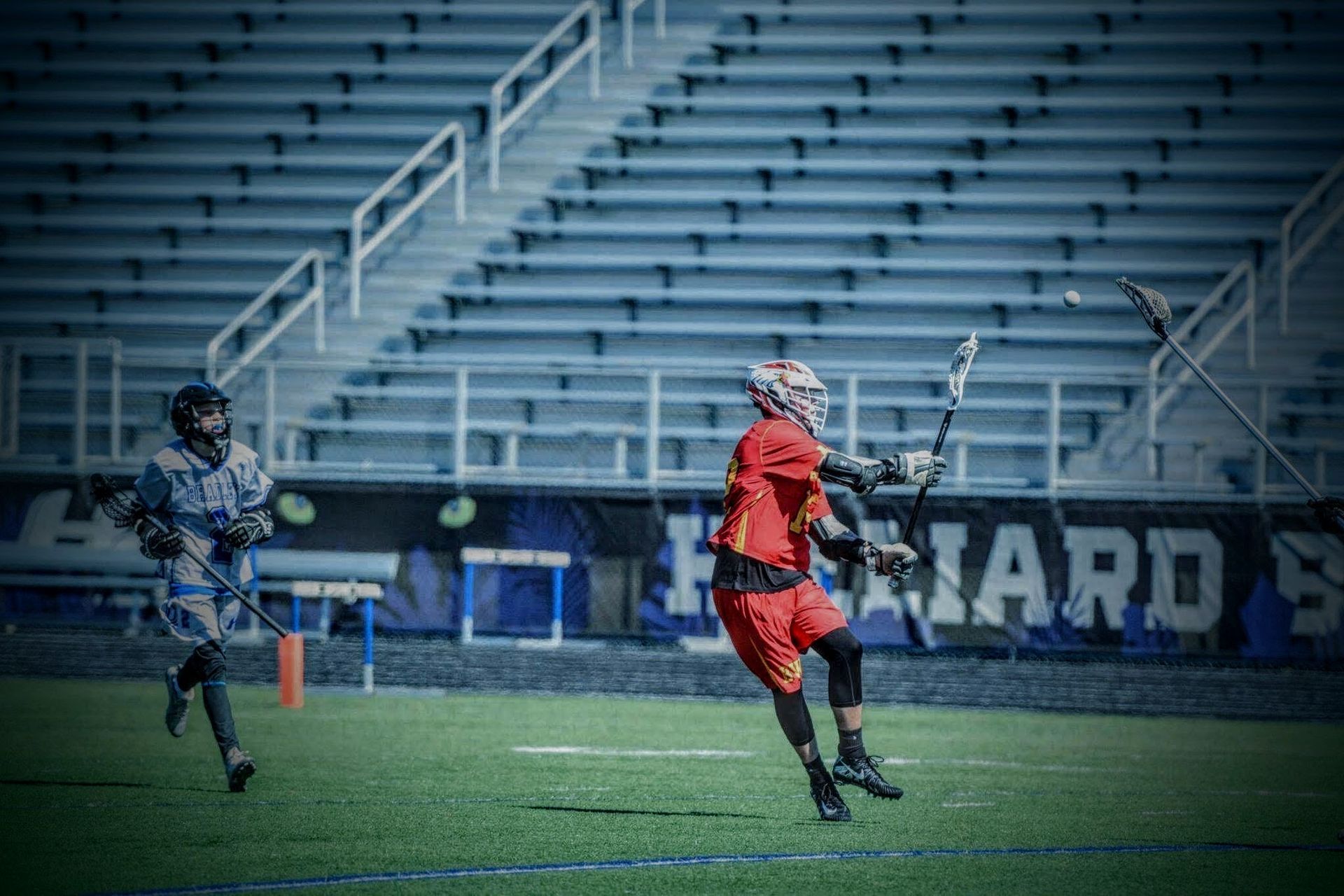 Two lacrosse players in red and gray run on a field with empty bleachers behind them