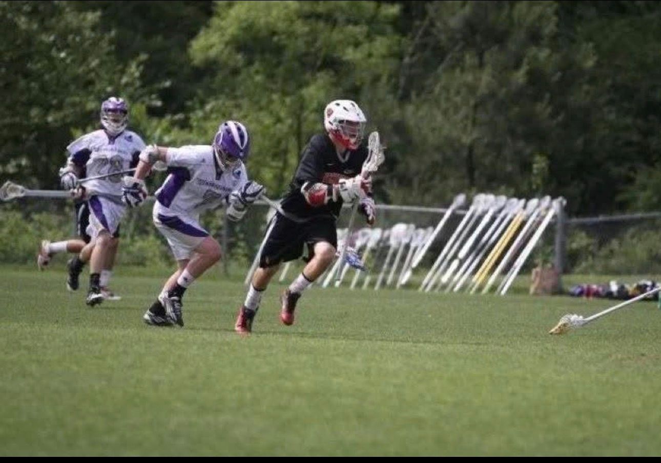 Lacrosse players sprint across a grassy field, with a black-clad player carrying the ball past two purple-uniformed opponents