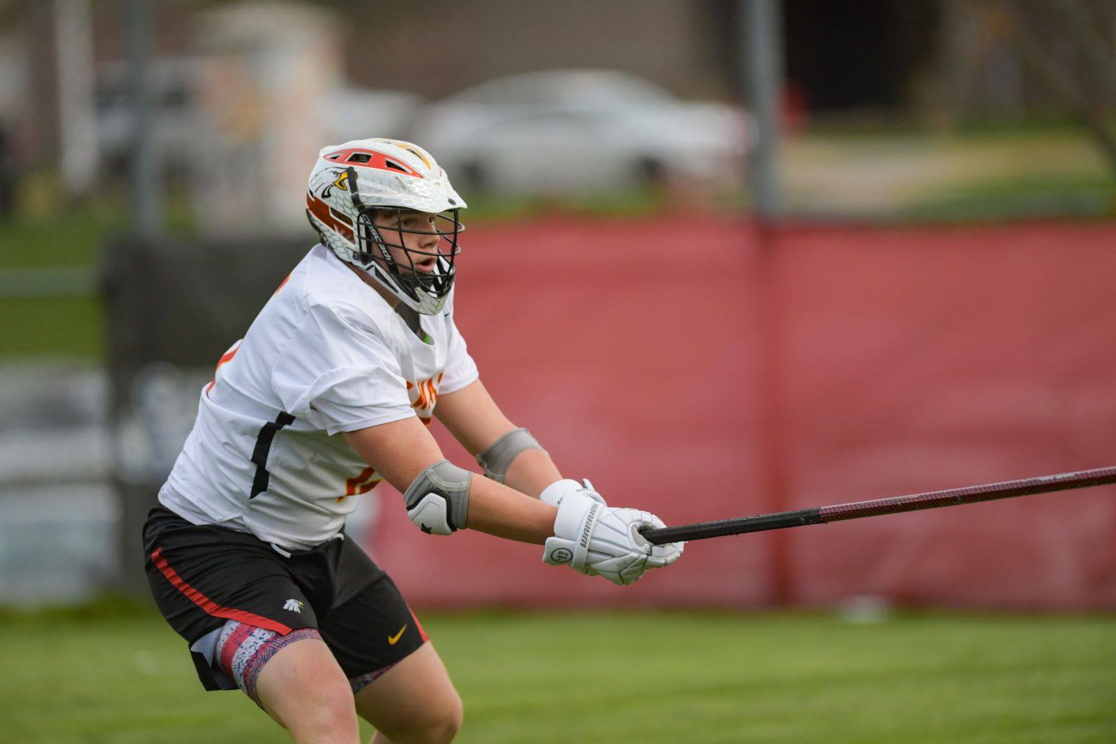 Lacrosse player in white jersey crouches on field, holding stick with a ball in the net.