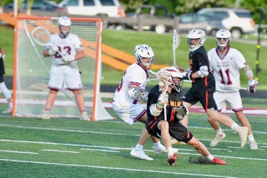 Lacrosse players in white and black uniforms battle for the ball near the goal on a green field.