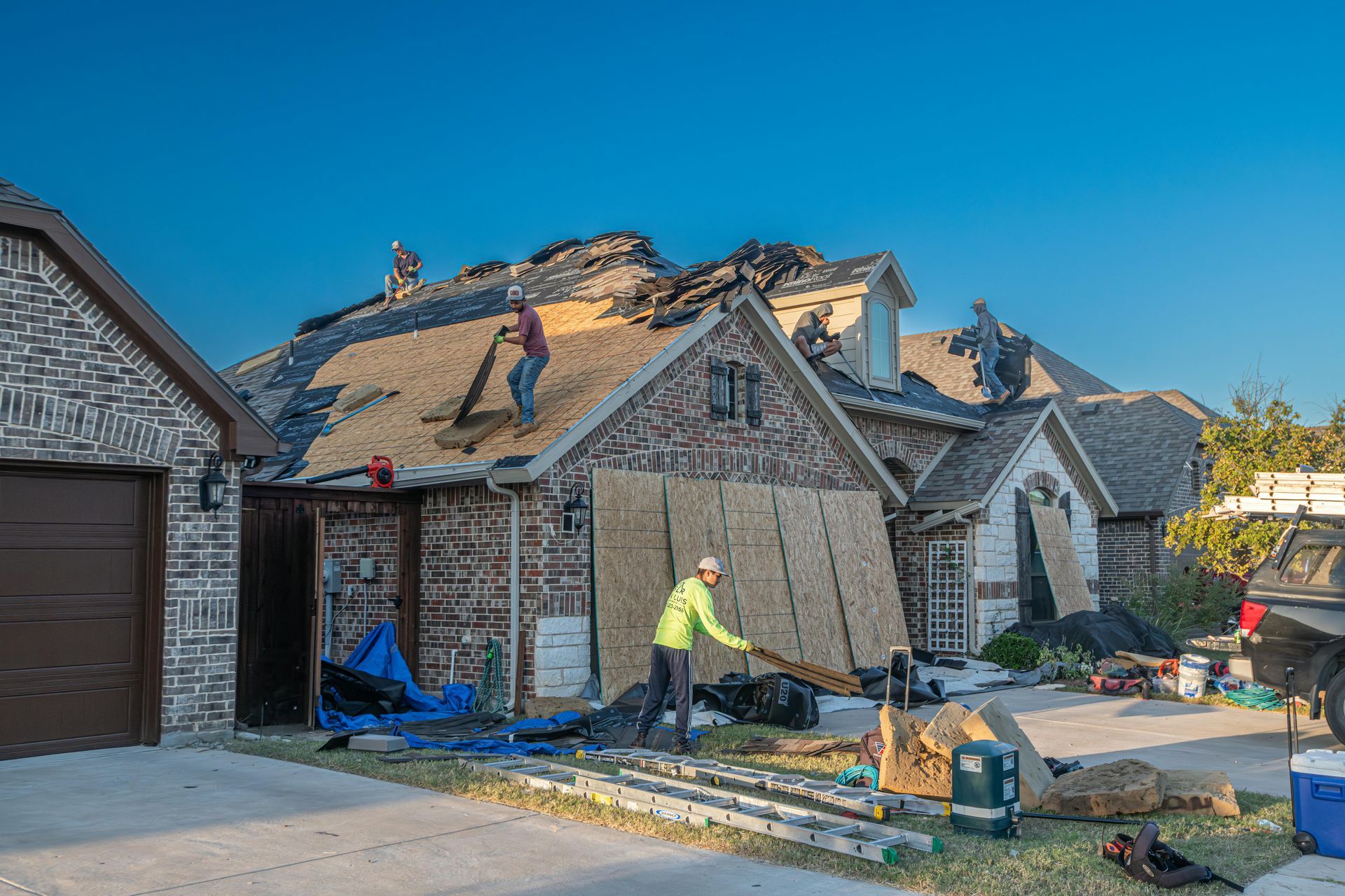 Workers repairing a damaged roof on a brick house; tools and materials scattered below.