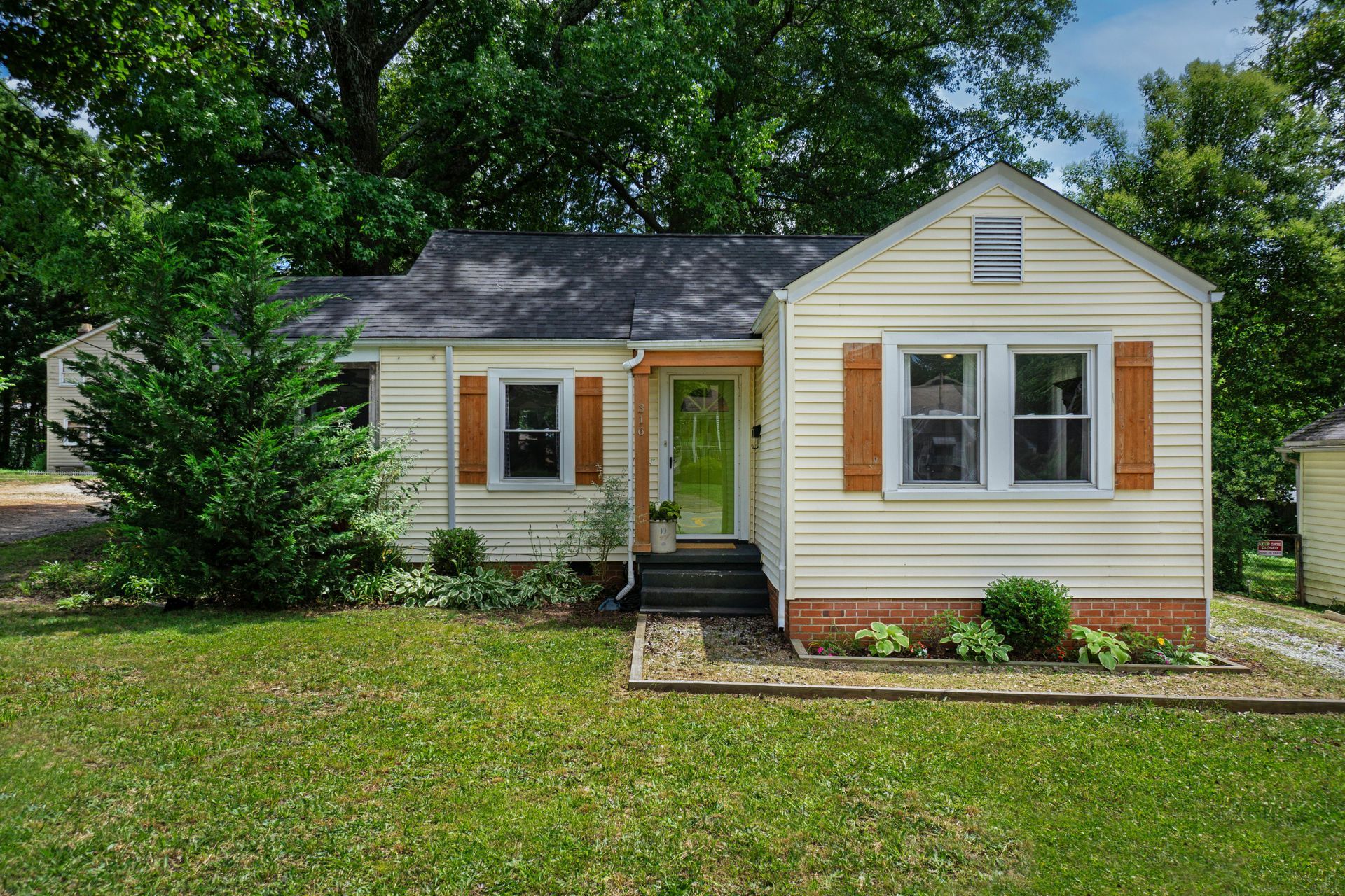Beige bungalow with brown shutters and a green door. Green lawn and trees surround the house.